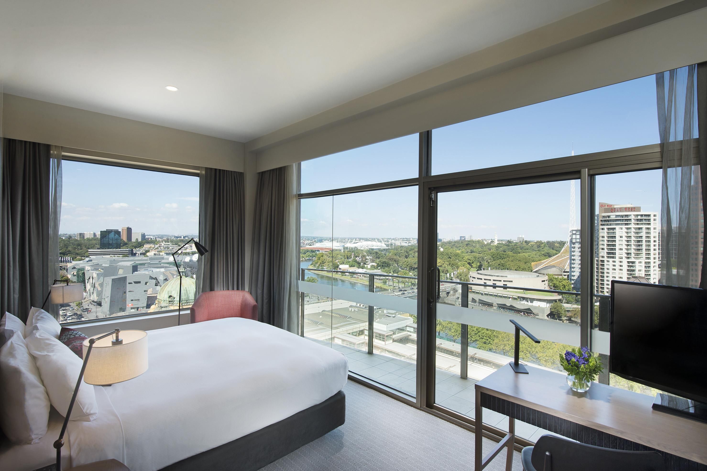 A hotel room with a large bed, a desk with a TV, and a view of a cityscape through large windows.