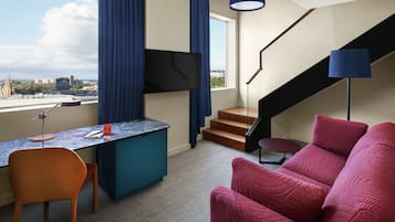 A modern hotel room with a desk, chair, and a sofa. There is a TV mounted on the wall, a staircase, and a view of the cityscape.