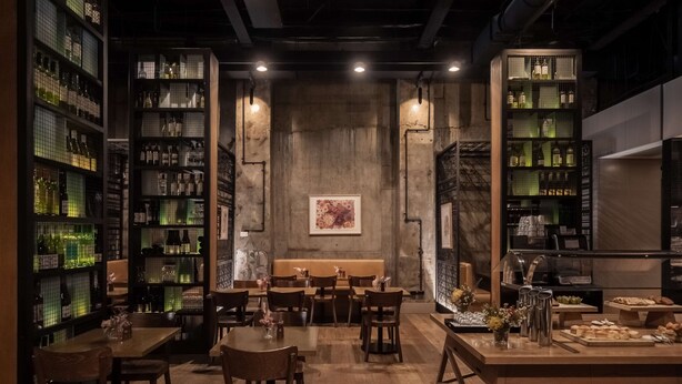 A restaurant interior with wooden tables and chairs, a central seating area, and shelves with bottles.