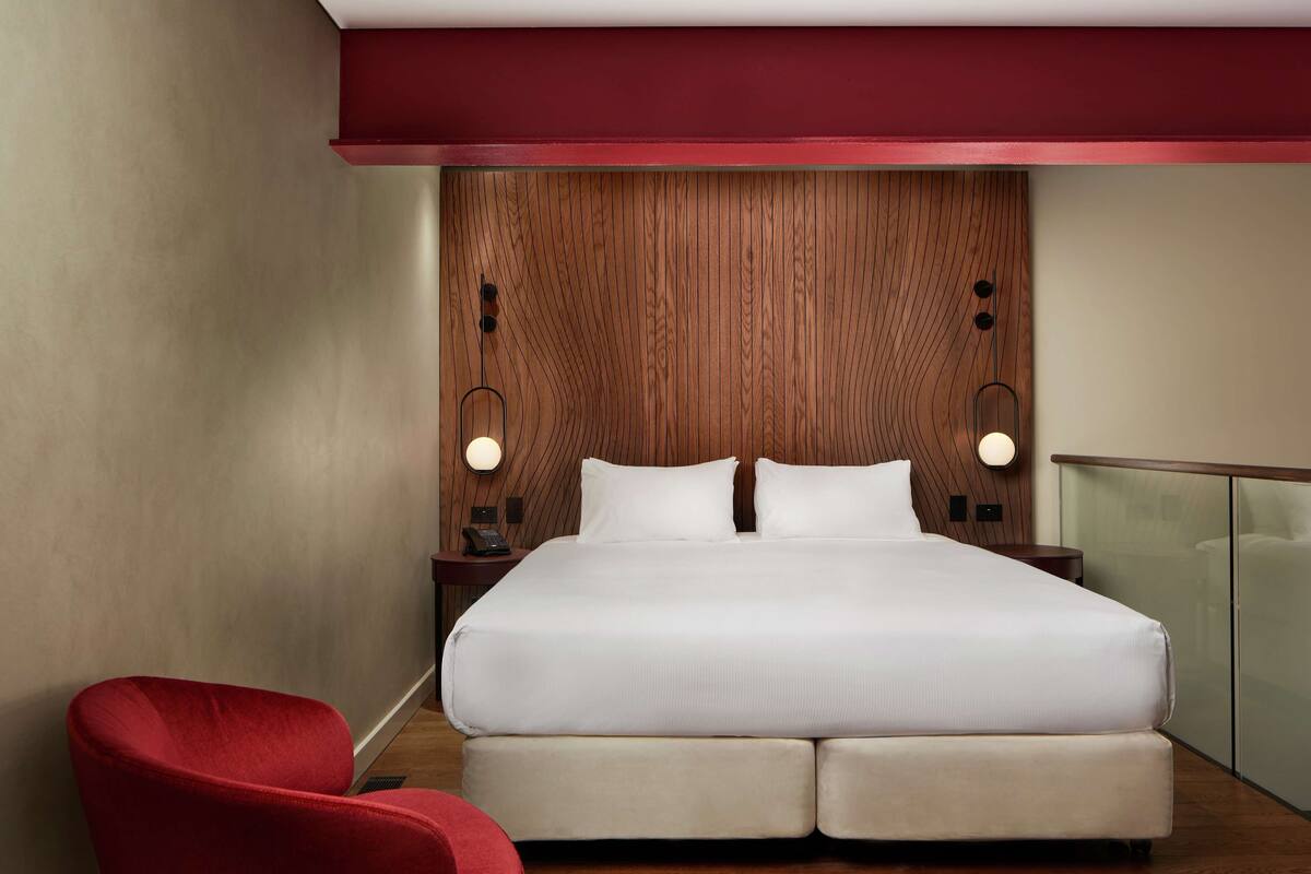 A modern hotel room with a large bed, wooden panel wall, two bedside lamps, and a red chair.