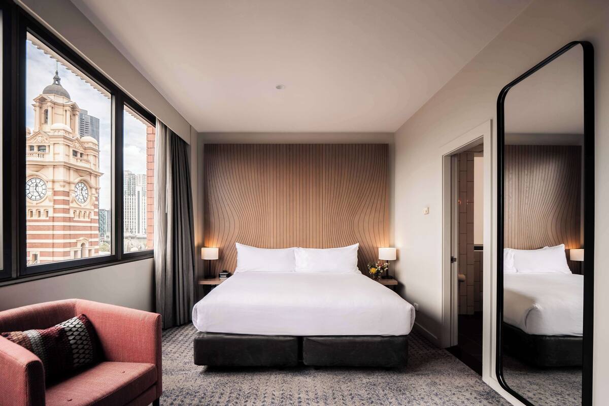 A hotel room with a large bed, two bedside tables with lamps, a large mirror, a red armchair, and a view of a historic building through the window.