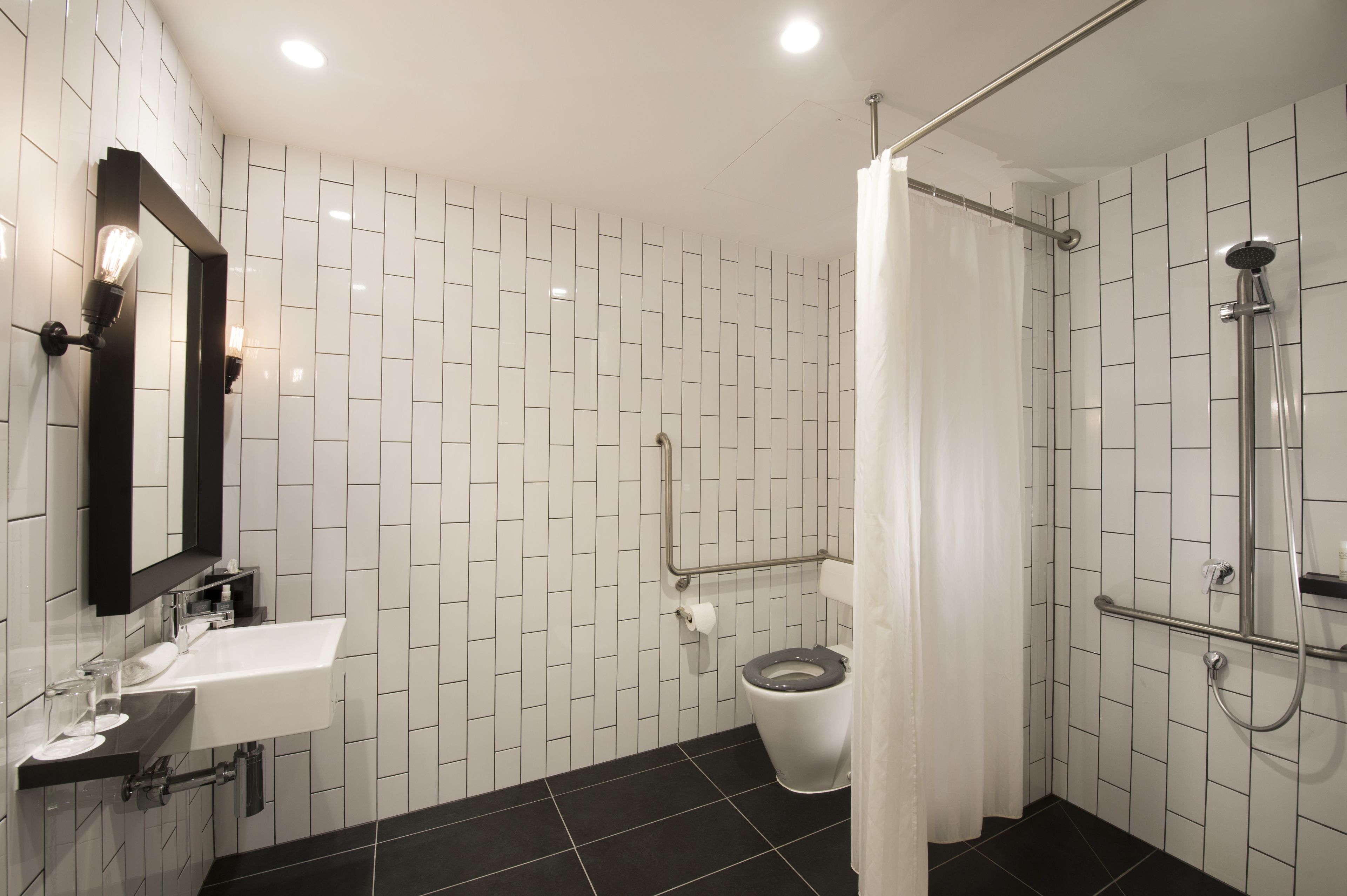 A modern bathroom with a white tiled wall, a black floor, a white sink with a mirror, a toilet, and a shower area.