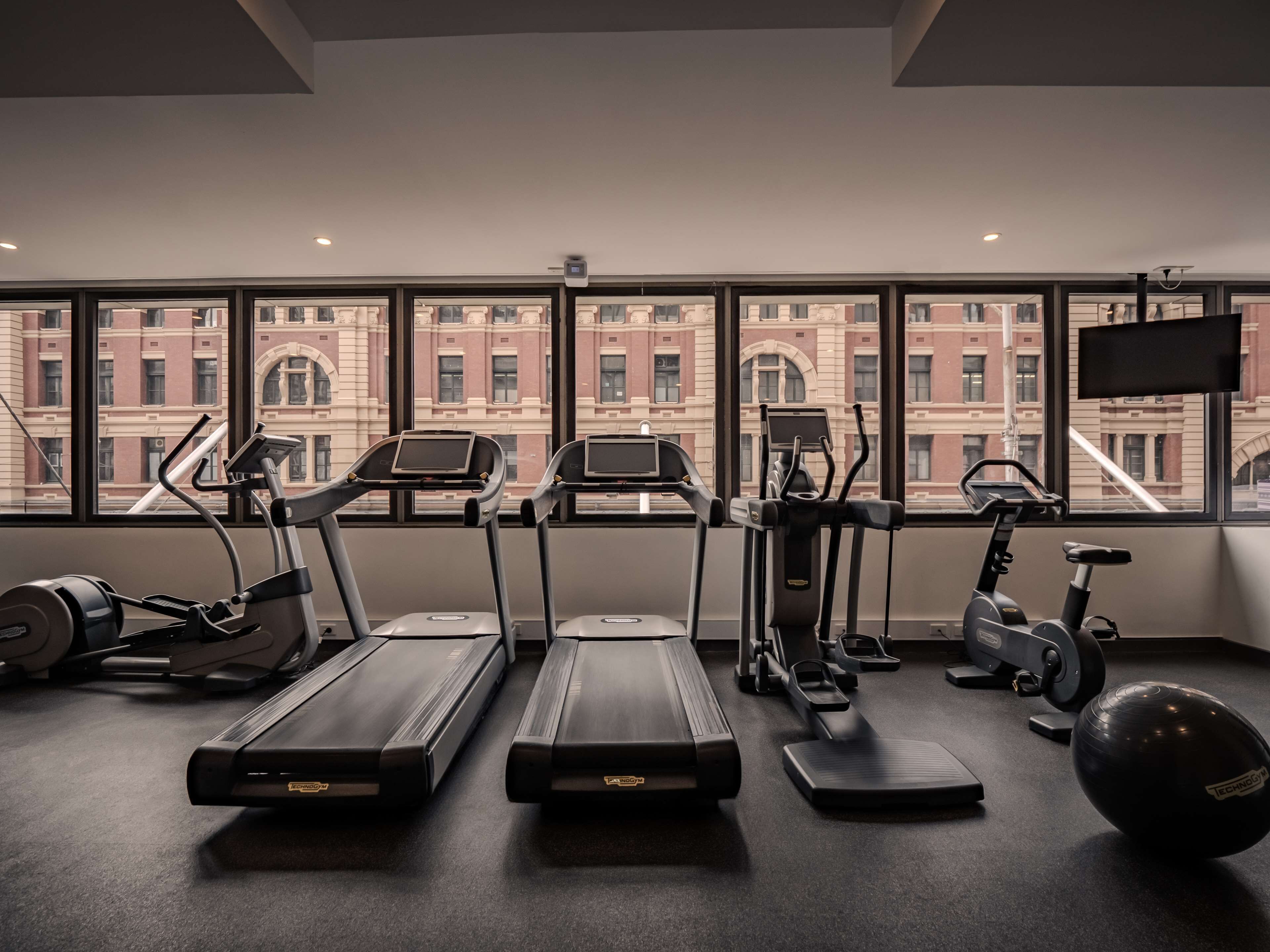 A gym with treadmills, elliptical machines, and a stationary bike, with a cityscape view through large windows.