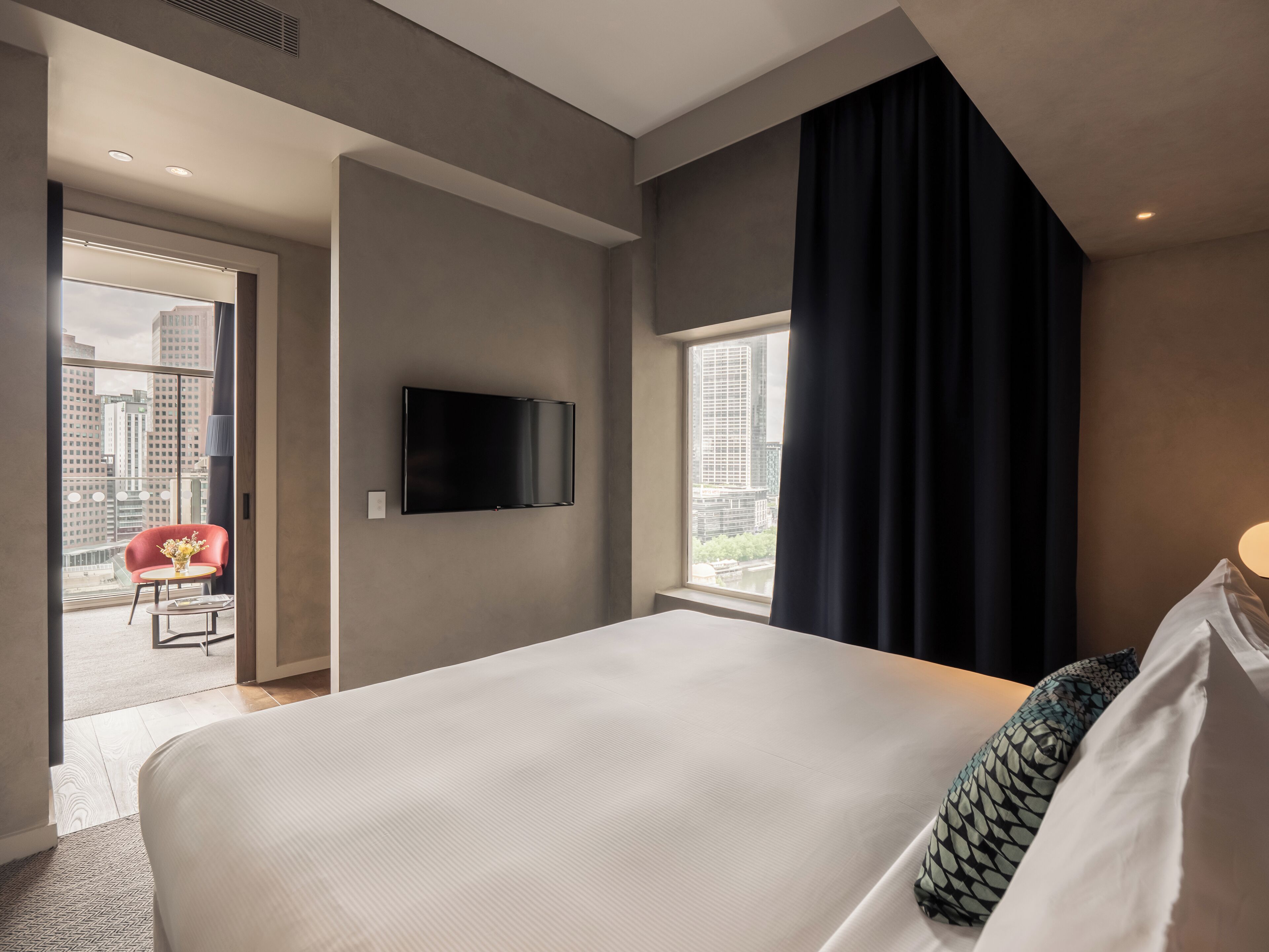 Un cuarto de hotel con una cama grande, un televisor de pantalla plana montado en la pared, una silla roja y vista a la ciudad por una ventana.