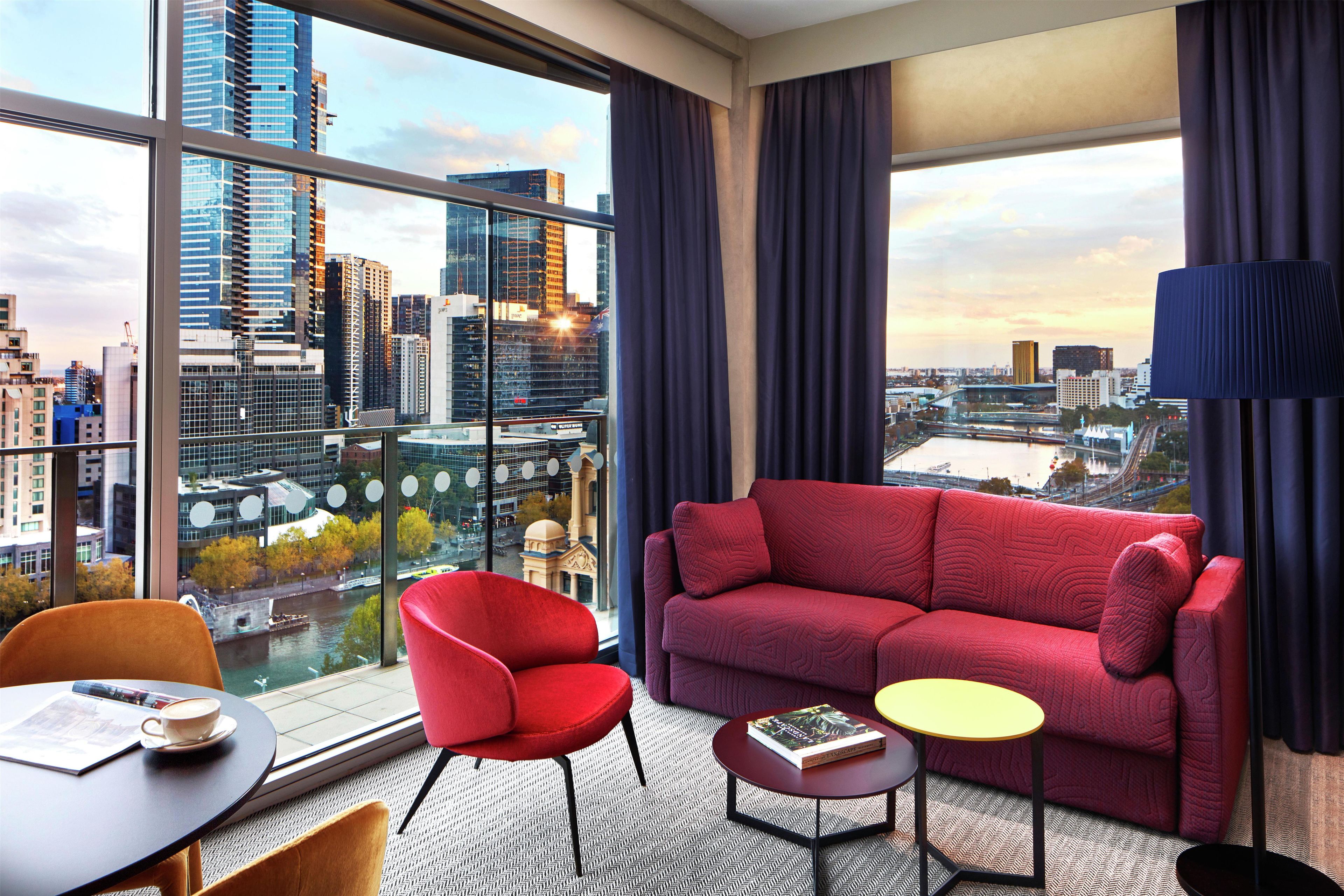 A modern hotel room with a red sofa, a small round table, and a chair. A large window offers a city view.
