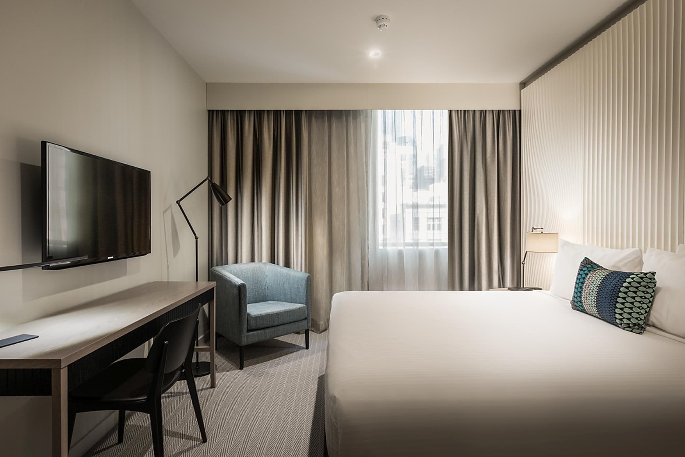 Habitación de hotel con cama, escritorio, silla, televisor, ventana con cortinas y lámpara.