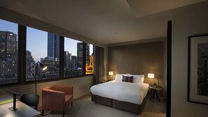A modern hotel room with a large bed, a desk, a chair, and a view of the city skyline through large windows.