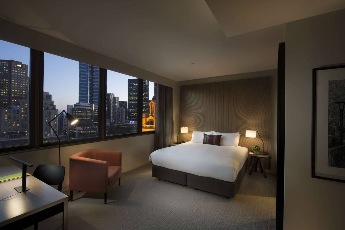A modern hotel room with a large bed, a desk, a chair, and a view of the city skyline through large windows.