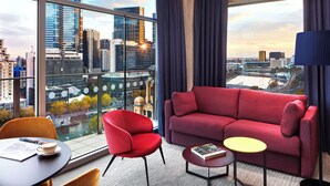 A modern hotel room with a large window offering a city view, a red sofa, a small round table, and a red armchair.