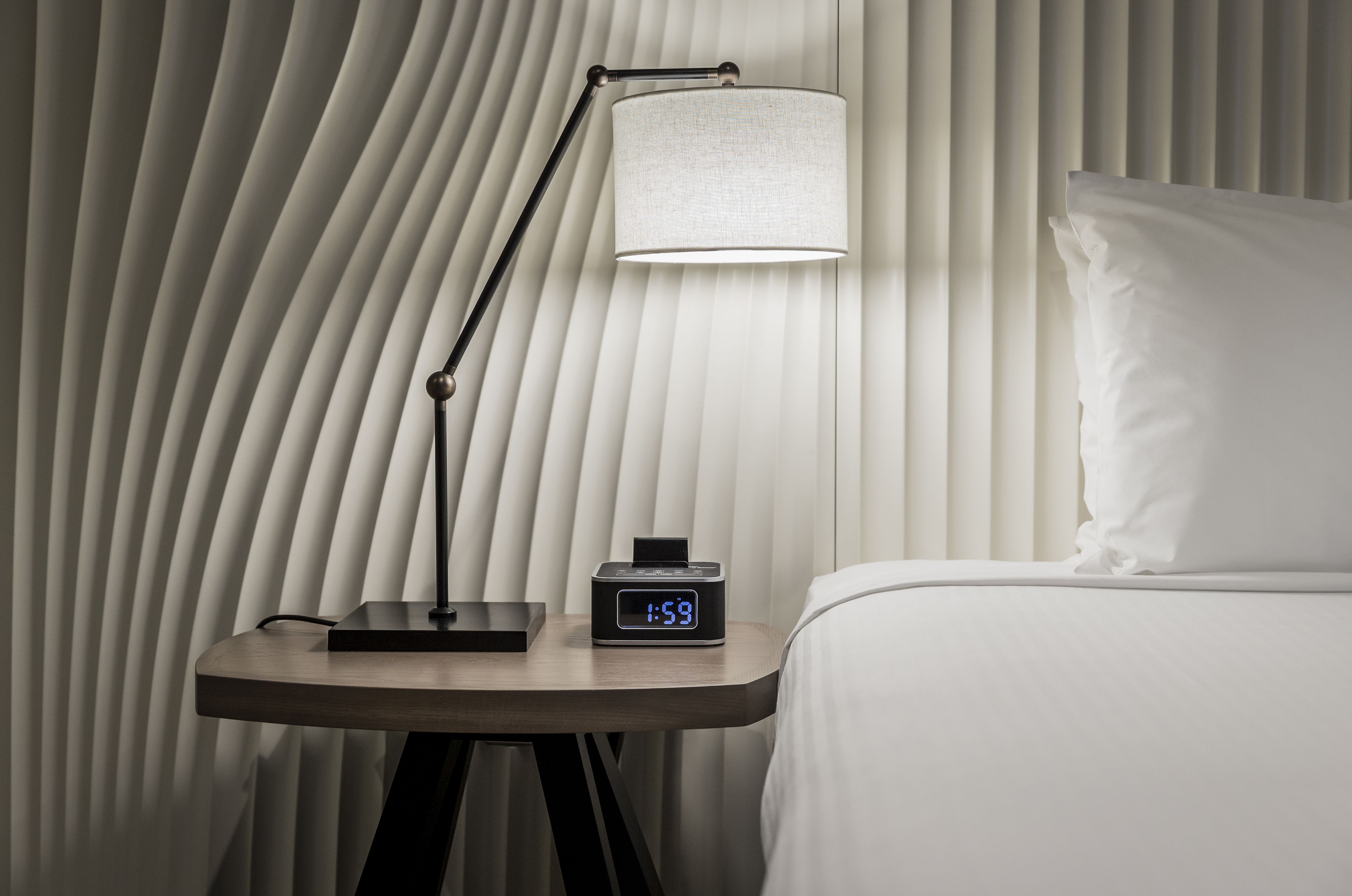 A bedside table with a digital clock displaying 1:59, a lamp, and a pillow against a wall with vertical blinds.
