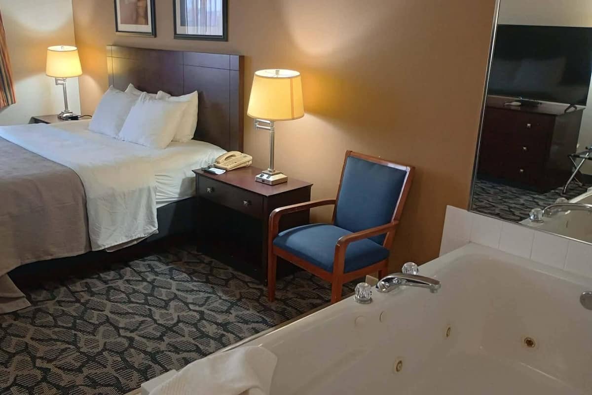 A hotel room with a bed, bedside lamps, a chair, a telephone, a mirror, and a bathtub.