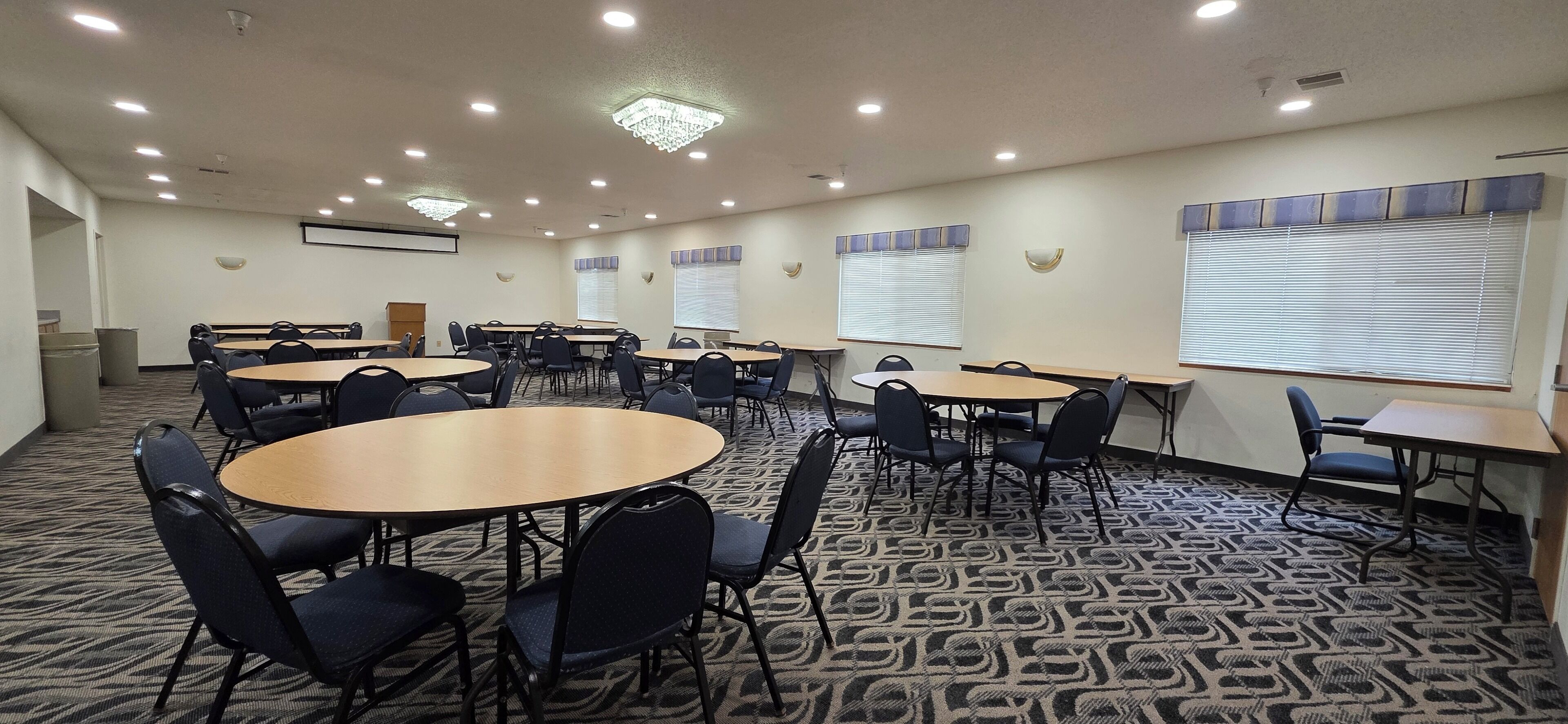 Un salón de conferencias con varias mesas y sillas, piso alfombrado, luces de techo y ventanas con persianas.