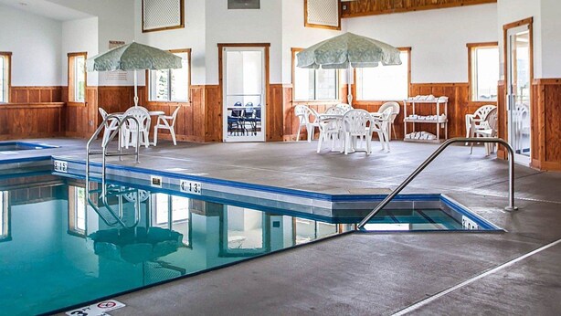 An indoor swimming pool with a diving board, surrounded by white plastic chairs and tables under green umbrellas. - Quality Inn & Suites Eldridge Davenport North (Eldridge)
