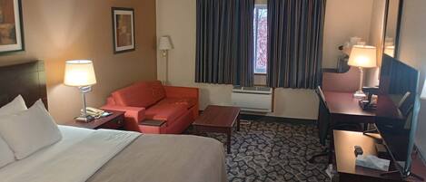 Una habitación de hotel con una cama, un sofá rojo, un escritorio con una lámpara y una ventana con cortinas.