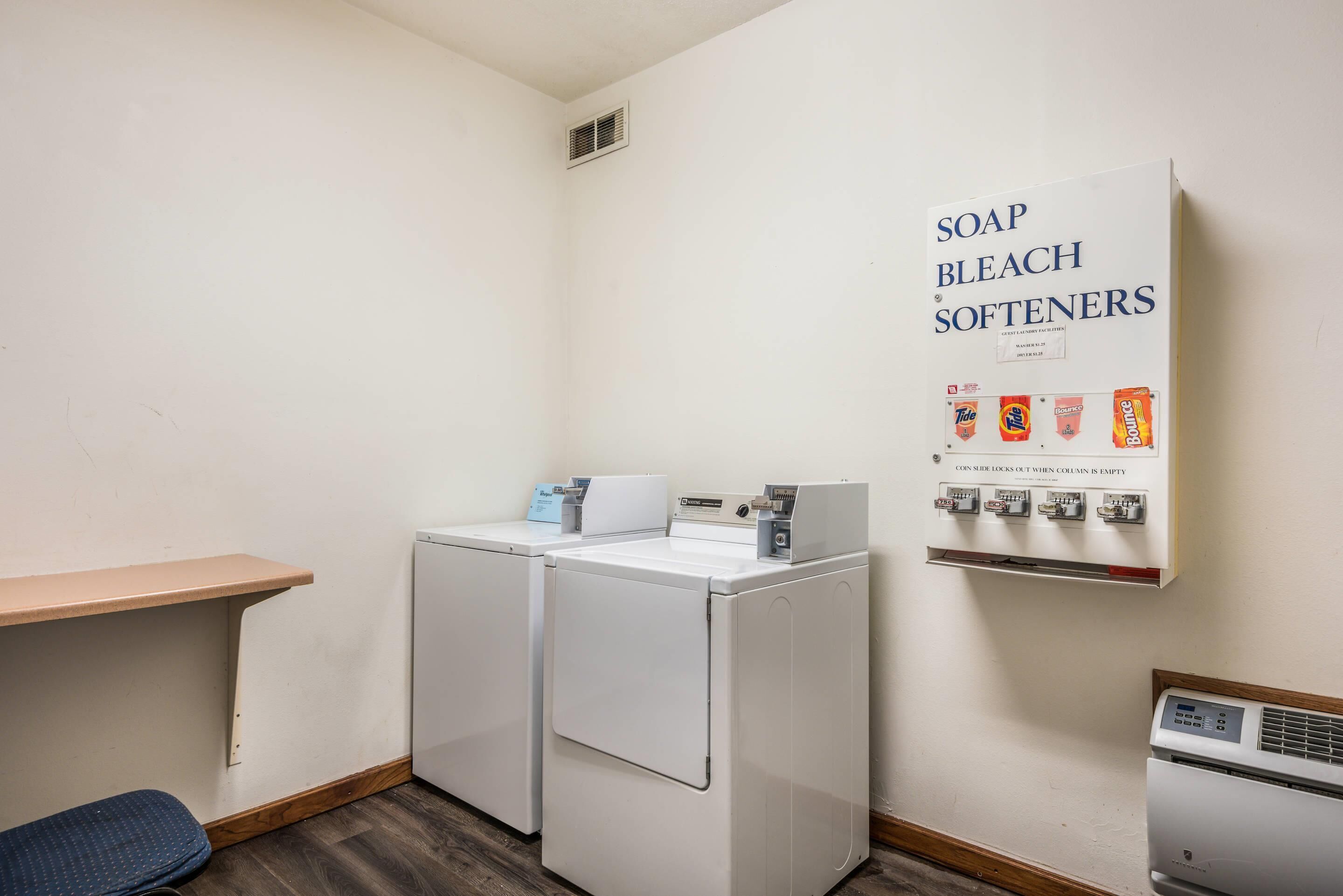 A laundry room with two white washing machines, a shelf with a blue item, and a wall-mounted soap, bleach, and softener dispenser.