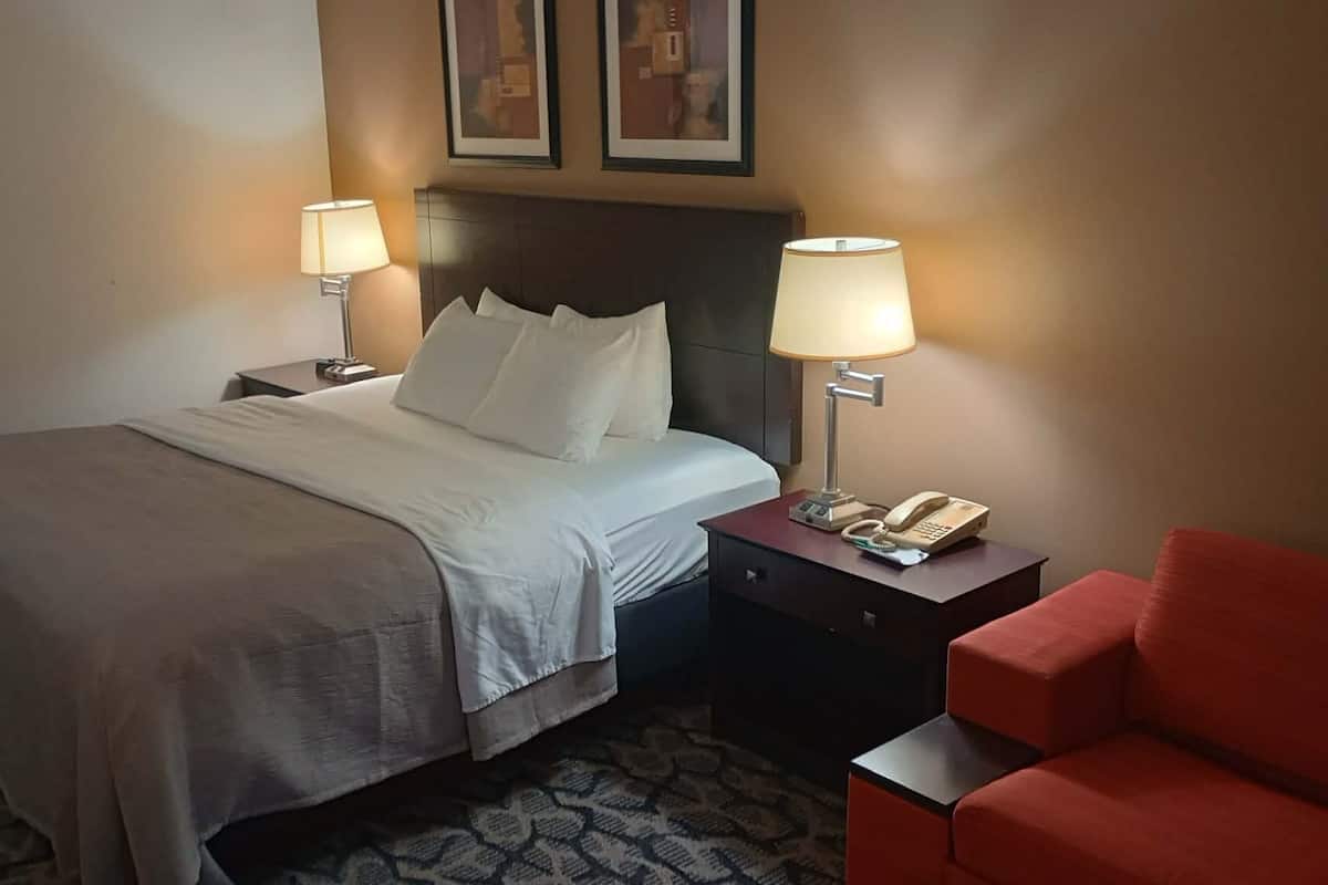 A hotel room with a bed, two bedside lamps, a telephone, a red chair, and a patterned carpet.