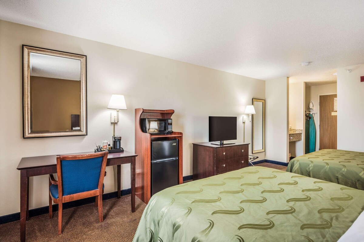 Een hotelkamer met twee bedden, een bureau met een stoel, een koffiezetapparaat, een televisie en een spiegel.