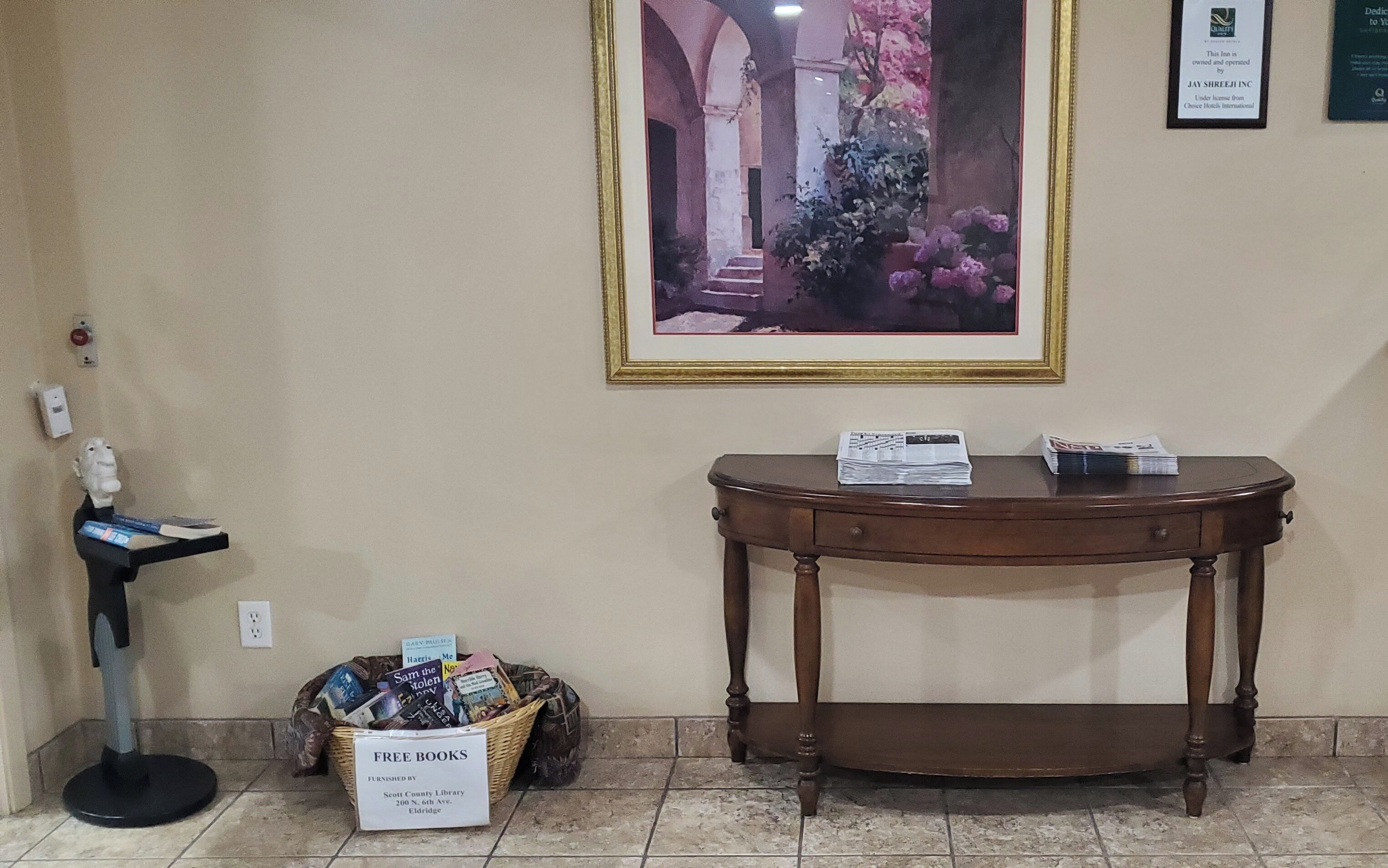 Un área de recepción con un cuadro enmarco, una mesa de madera, una cesta de libros gratuitos y una estatua.