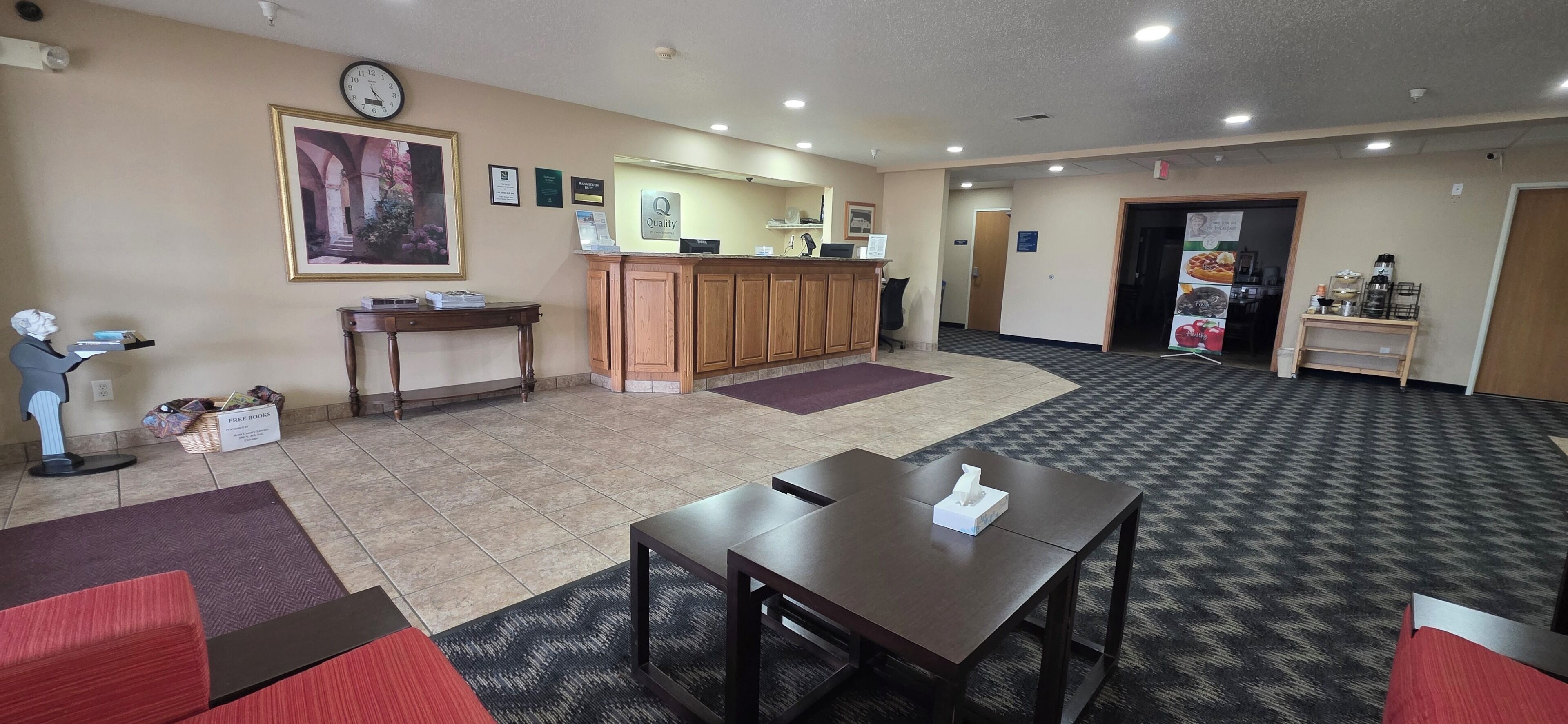 A hotel lobby with a reception desk, a clock, a framed picture, a coffee station, and a seating area.