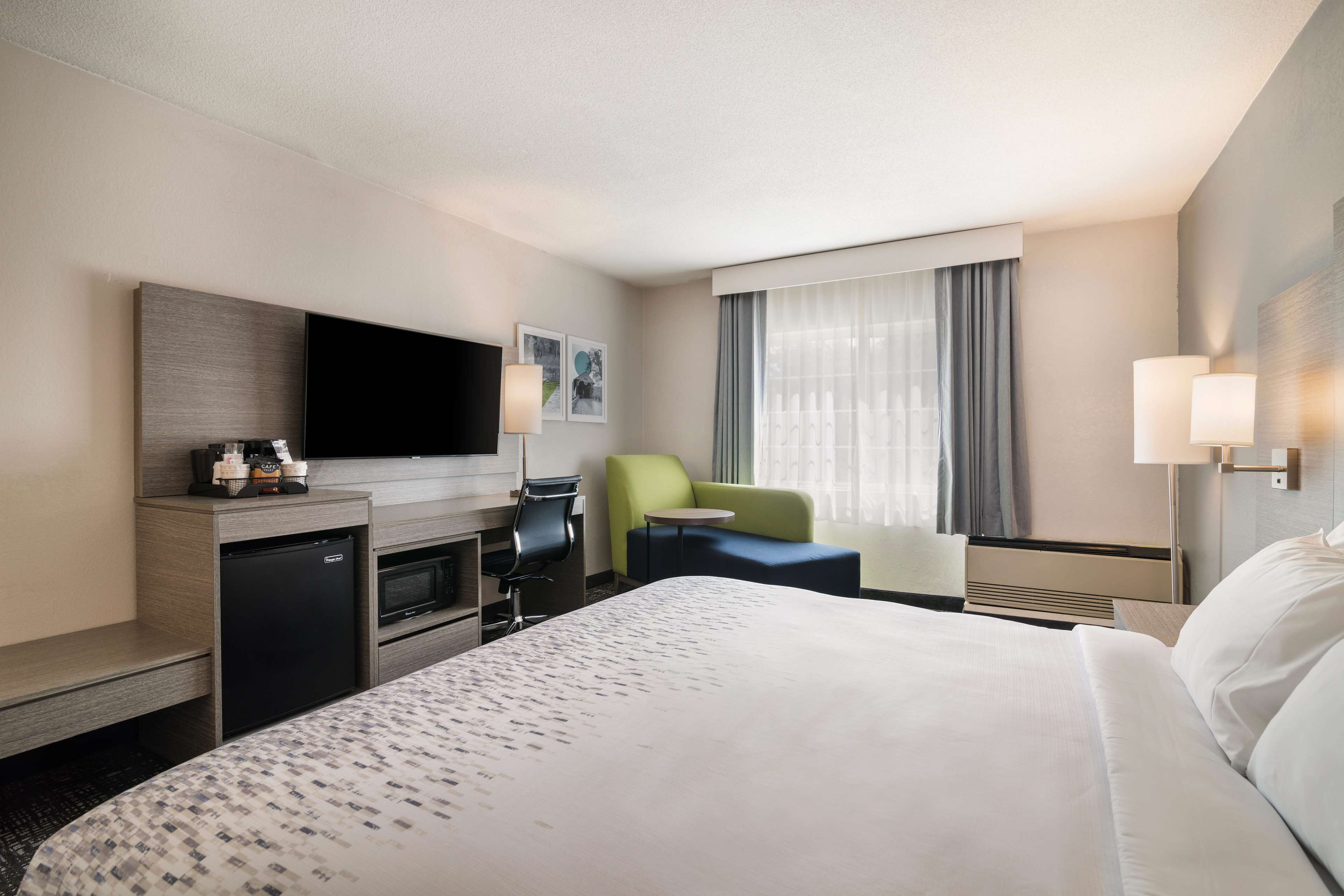 A hotel room with a bed, a desk with a flat-screen TV, a mini-fridge, a chair, and a window with curtains.