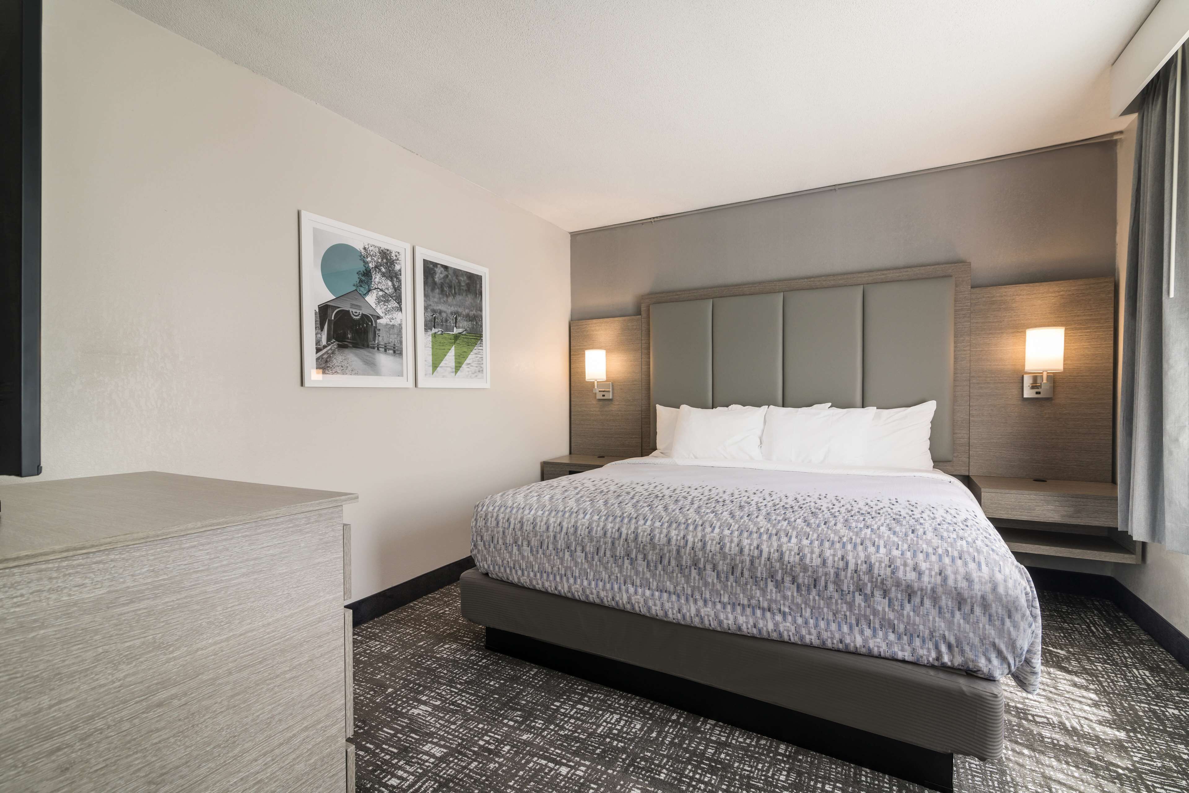 A hotel room with a large bed, a nightstand, a dresser, and two wall-mounted lamps.