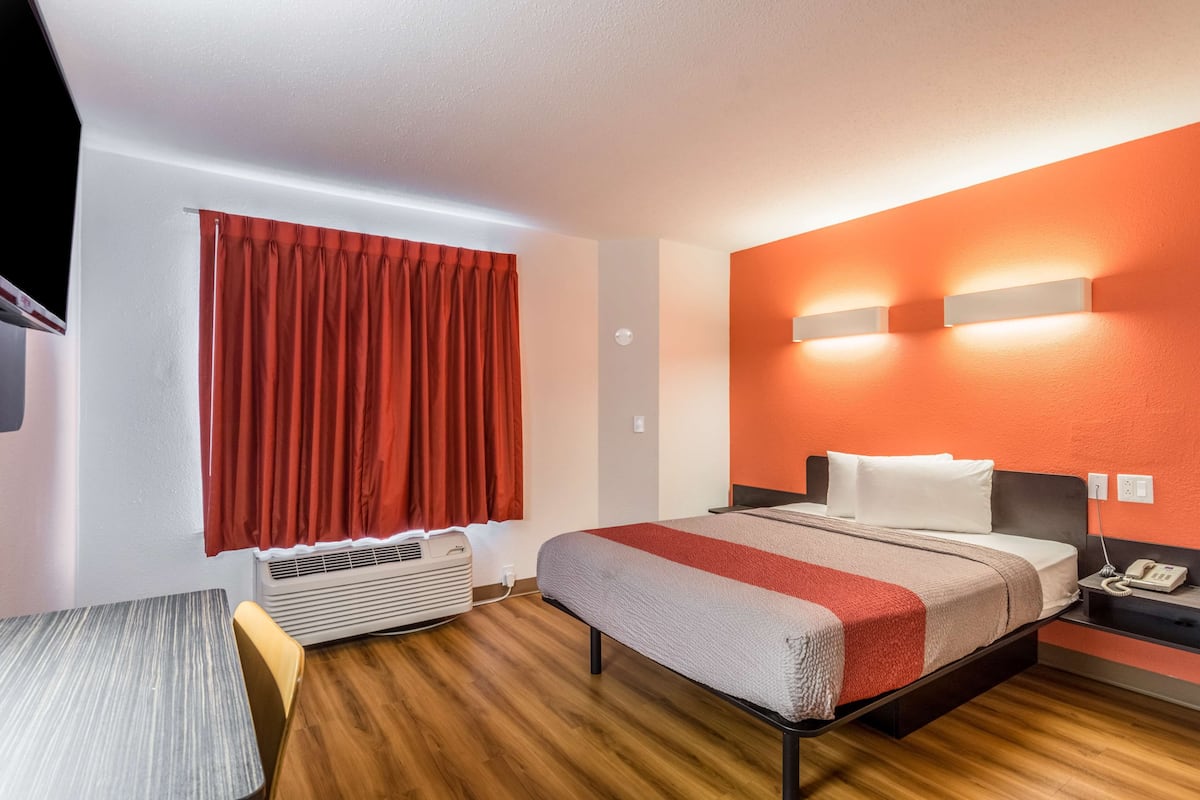 A hotel room with a bed, a desk, a chair, an air conditioner, and a telephone on the nightstand.
