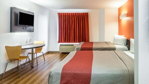 Una habitación de hotel con una televisión de pantalla plana montada en la pared, una mesa de comedor con sillas, una cama con colcha roja y gris, y una ventana con cortinas rojas.
