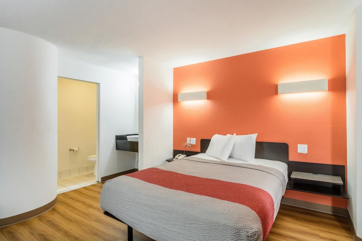 A hotel room with a bed, bedside tables, a nightstand, a wall-mounted light, and a door leading to a bathroom.