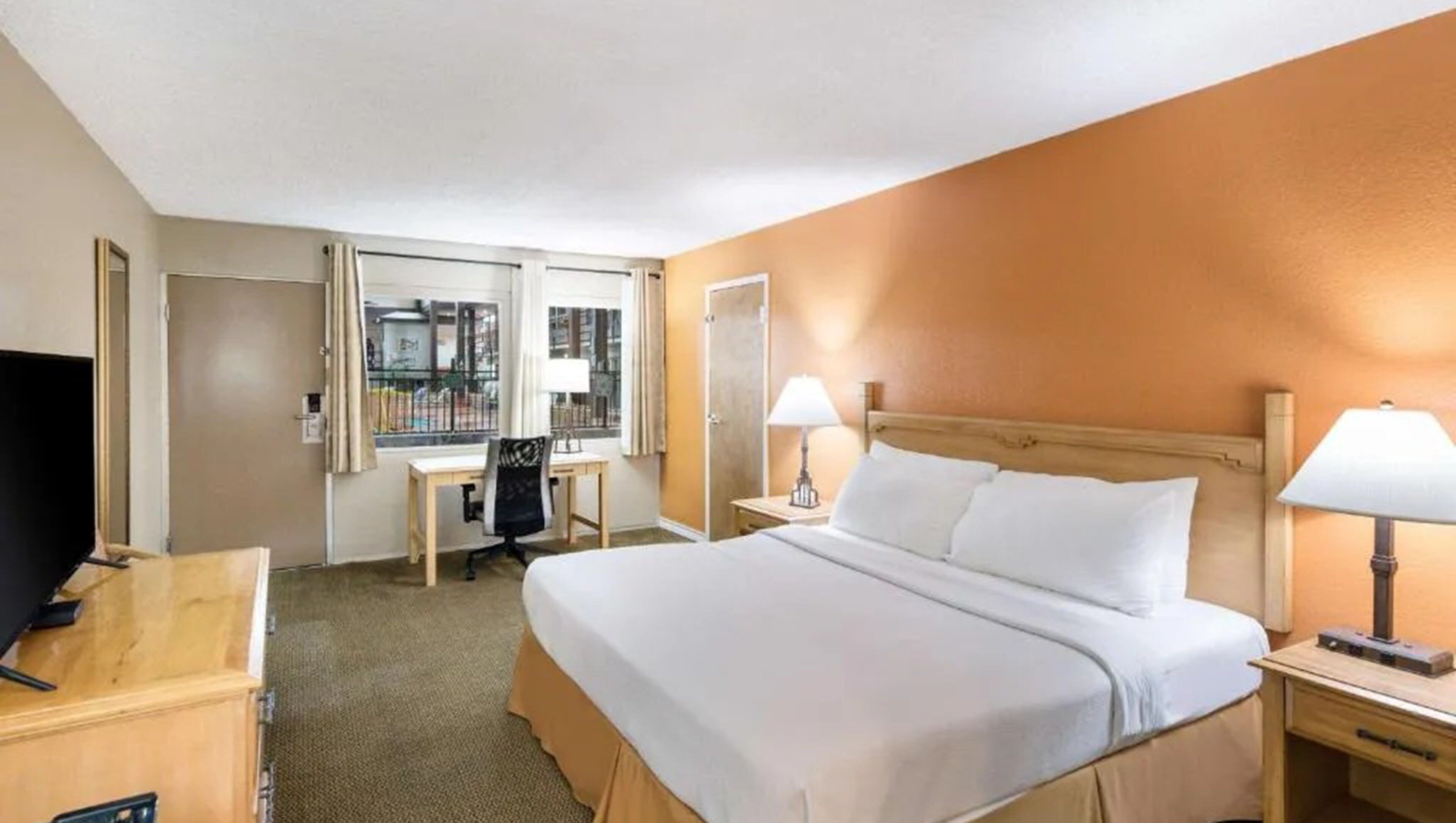 A hotel room with a large bed, two bedside tables with lamps, a desk with a chair, a TV, and a window with curtains.