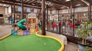 Un área de juegos para niños con tobogán y estructuras para escalar, rodeada por una zona vallada con sillas y mesas rojas.