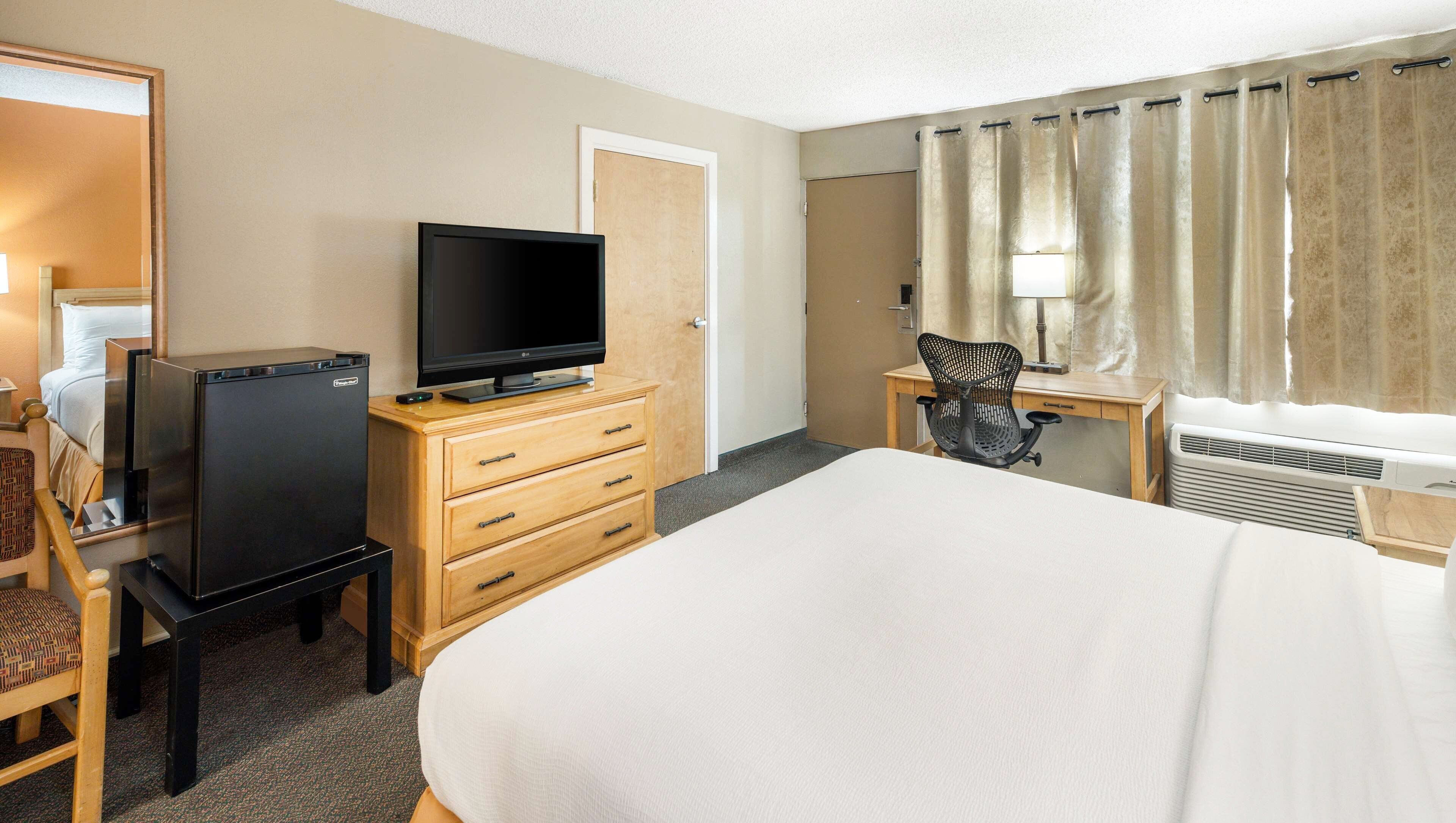 A hotel room with a bed, a desk with a chair, a television, a refrigerator, and a window with curtains.