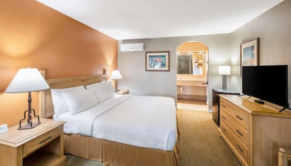 A hotel room with a bed, bedside lamps, a TV, a dresser, and a bathroom visible through an archway. - Pearl on the Concho (San Angelo)