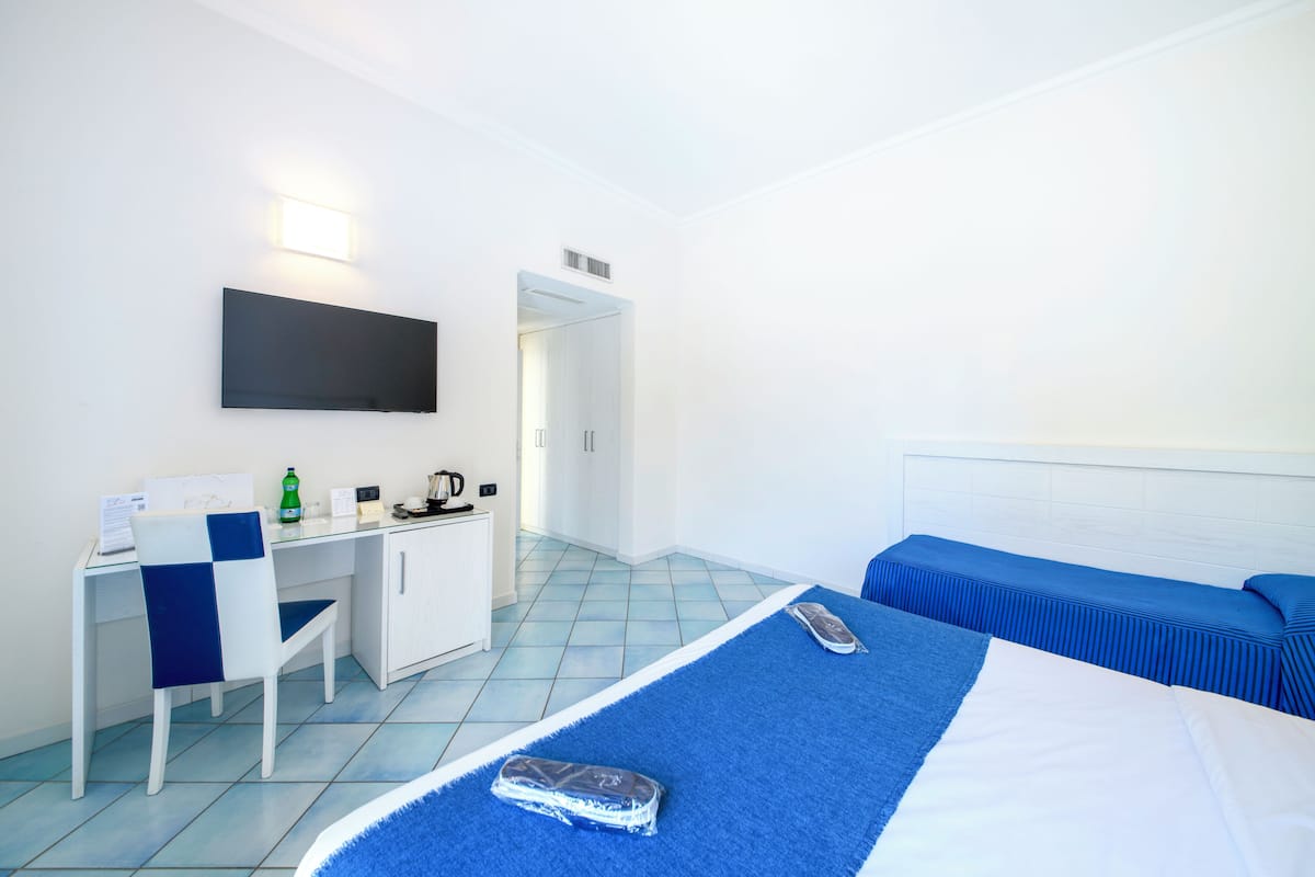 A hotel room with a bed, a desk with a chair, a television mounted on the wall, and a kitchenette area.