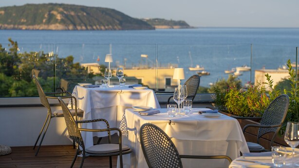 A dining area with tables set for two, overlooking a coastal view with boats and greenery. - Hotel Villa Durrueli Resort & Spa (Ischia)