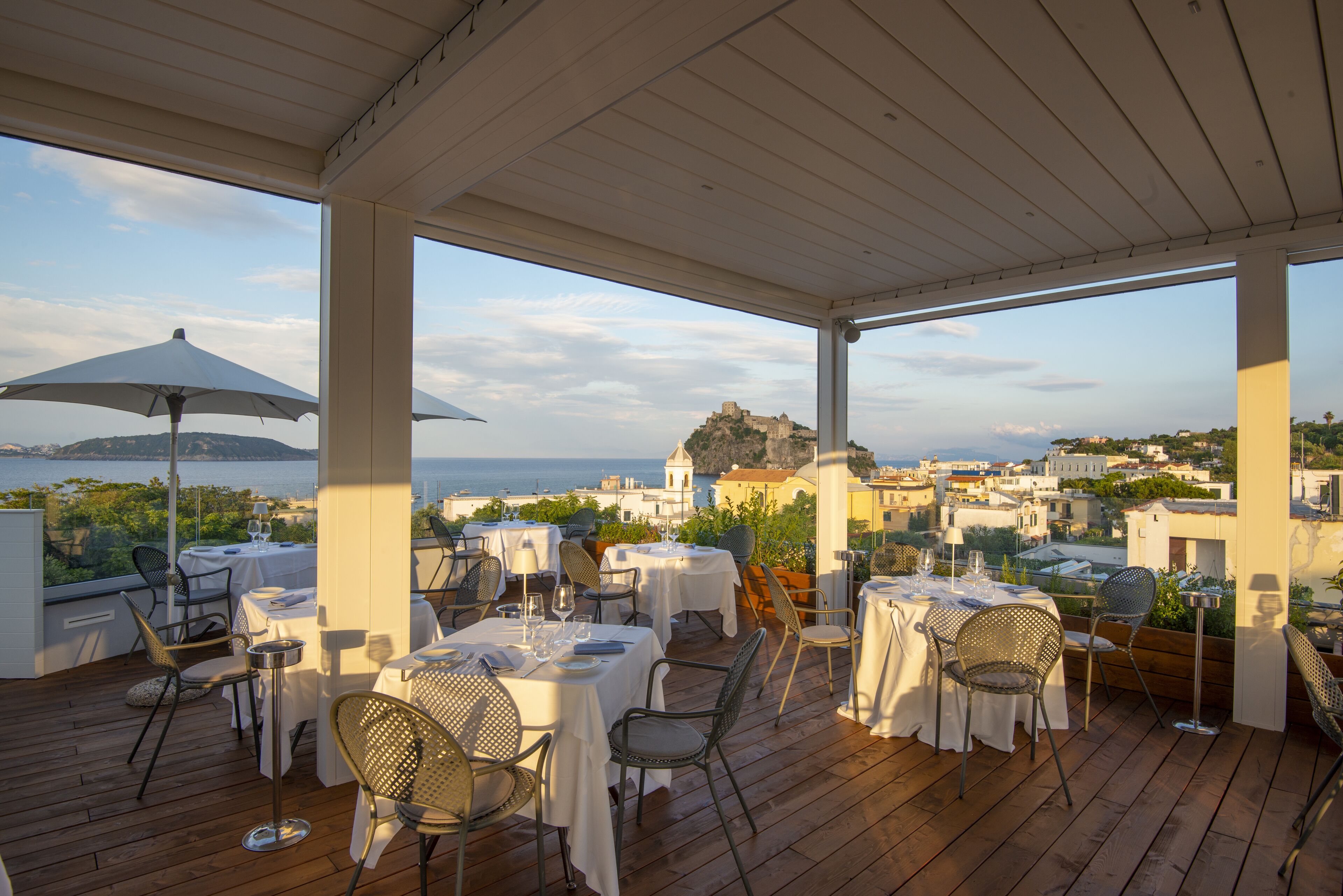 An outdoor dining area with tables and chairs, overlooking a scenic view of a coastal town and a hilltop castle.