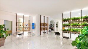 A spacious lobby with a high ceiling, white tiled floor, and a variety of seating options including chairs and a sofa. There are multiple shelves with potted plants and a large screen displaying an image. - Hotel Villa Durrueli Resort & Spa (Ischia)