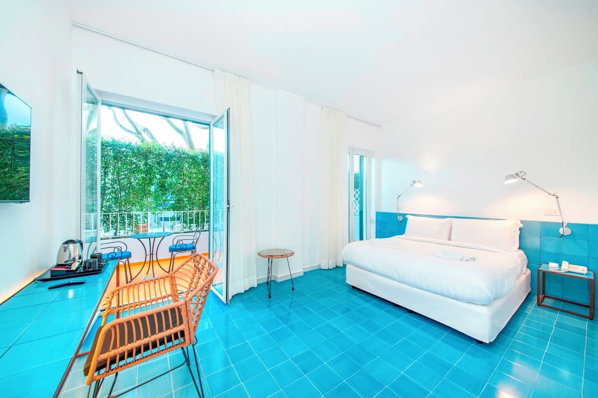 A hotel room with a large bed, a small table, a chair, and a balcony with a view of greenery.
