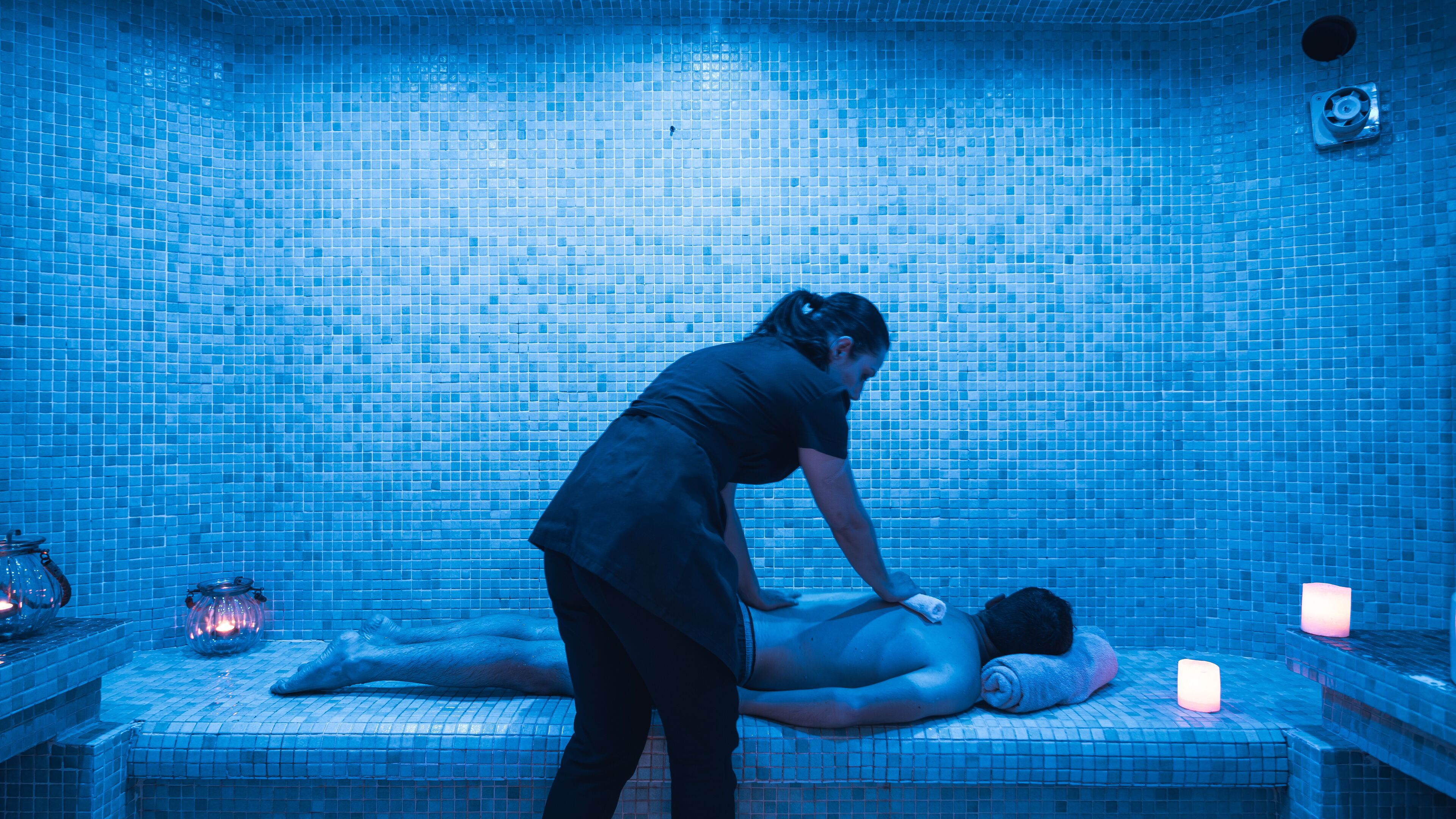 Una persona recibiendo un masaje en una habitación con pared azul de azulejos y una mesita con una vela.