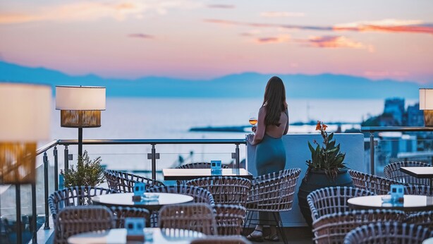 A terrace with wicker chairs and tables, overlooking a body of water and a cityscape during sunset. - Secret Paradise Hotel & Spa (Nea Propontida)