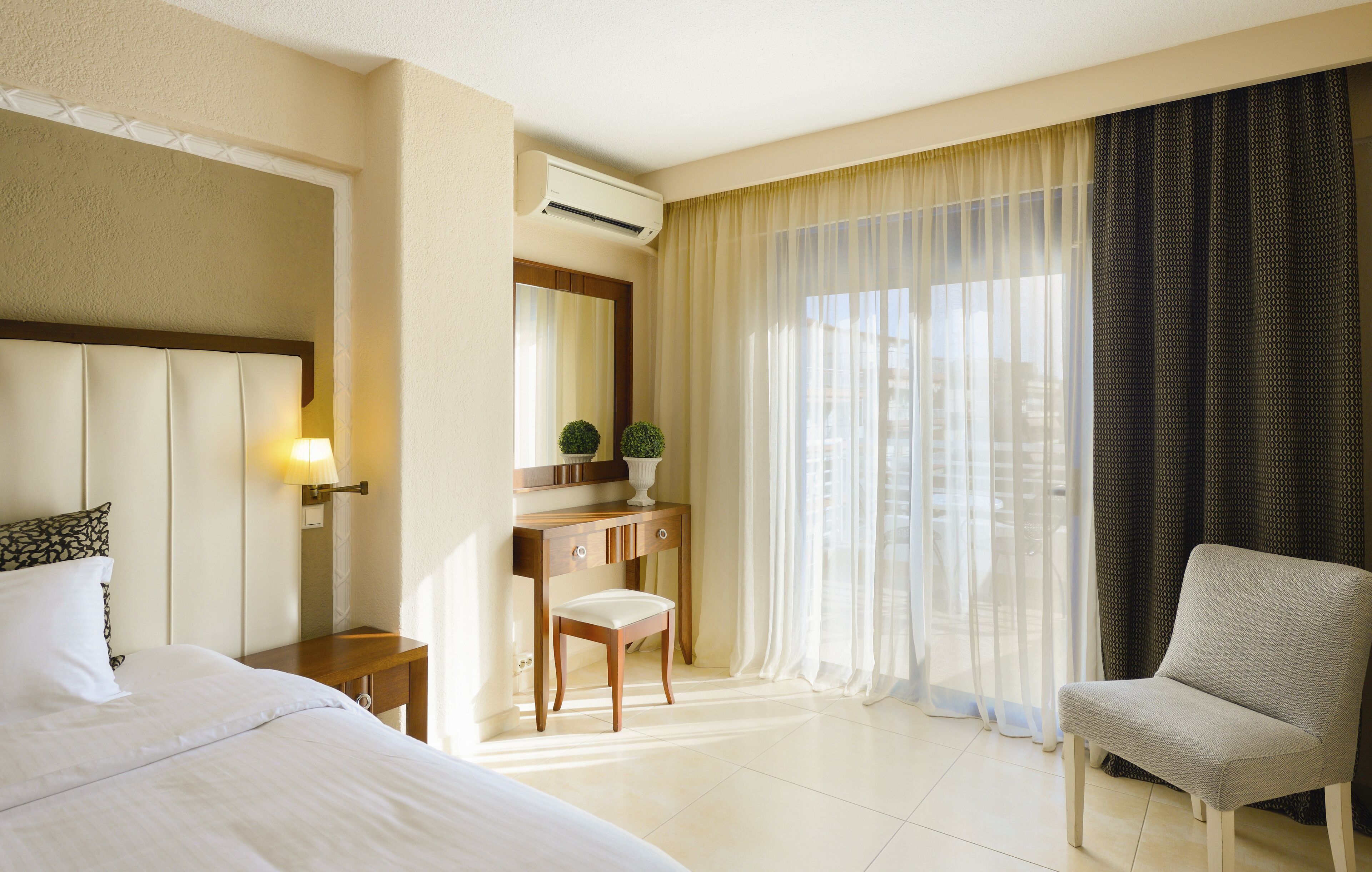 A hotel room with a bed, a desk with a chair, a mirror, and a window with curtains.
