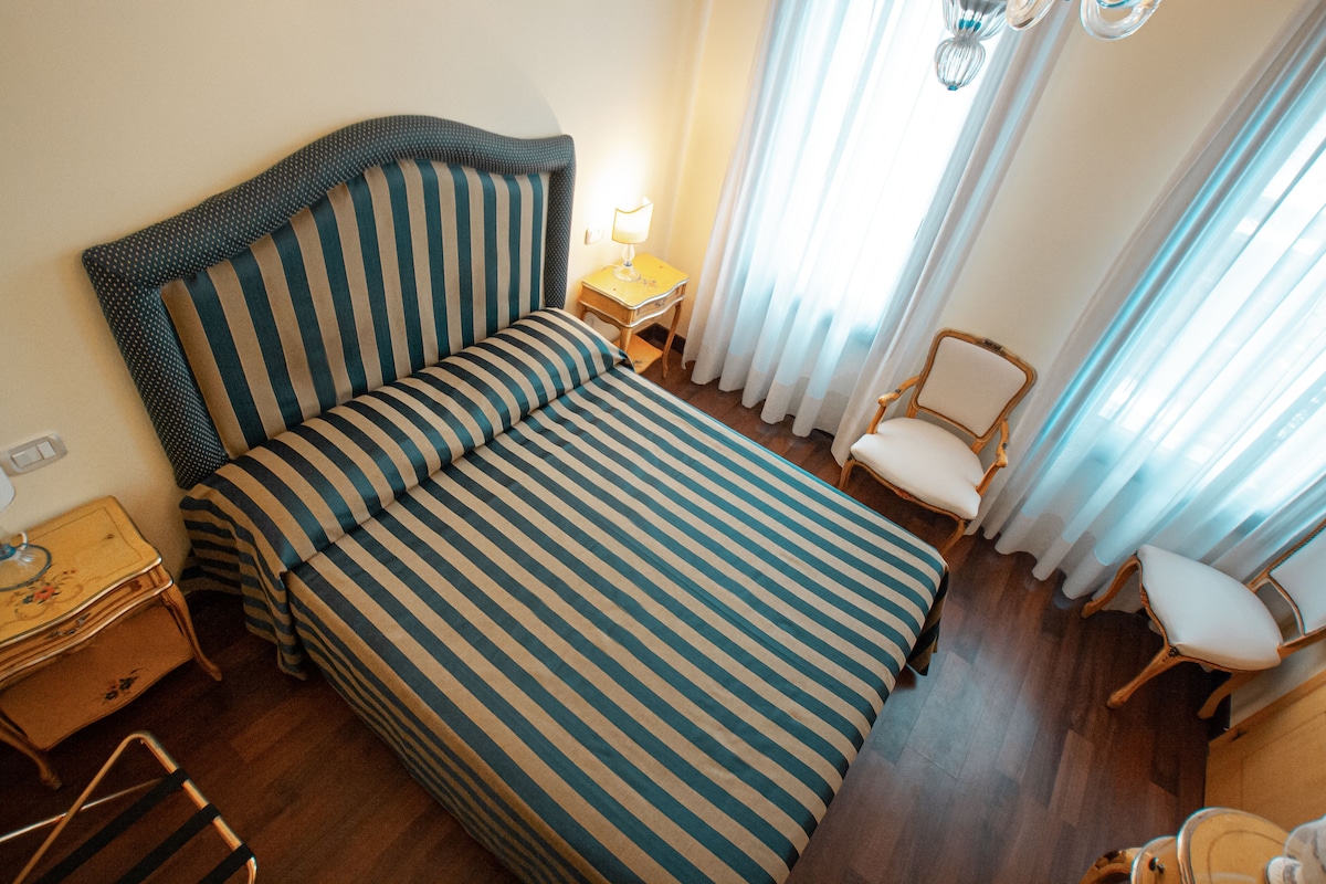 A hotel room with a large bed, a bedside table with a lamp, a chair, and a small table with a suitcase.