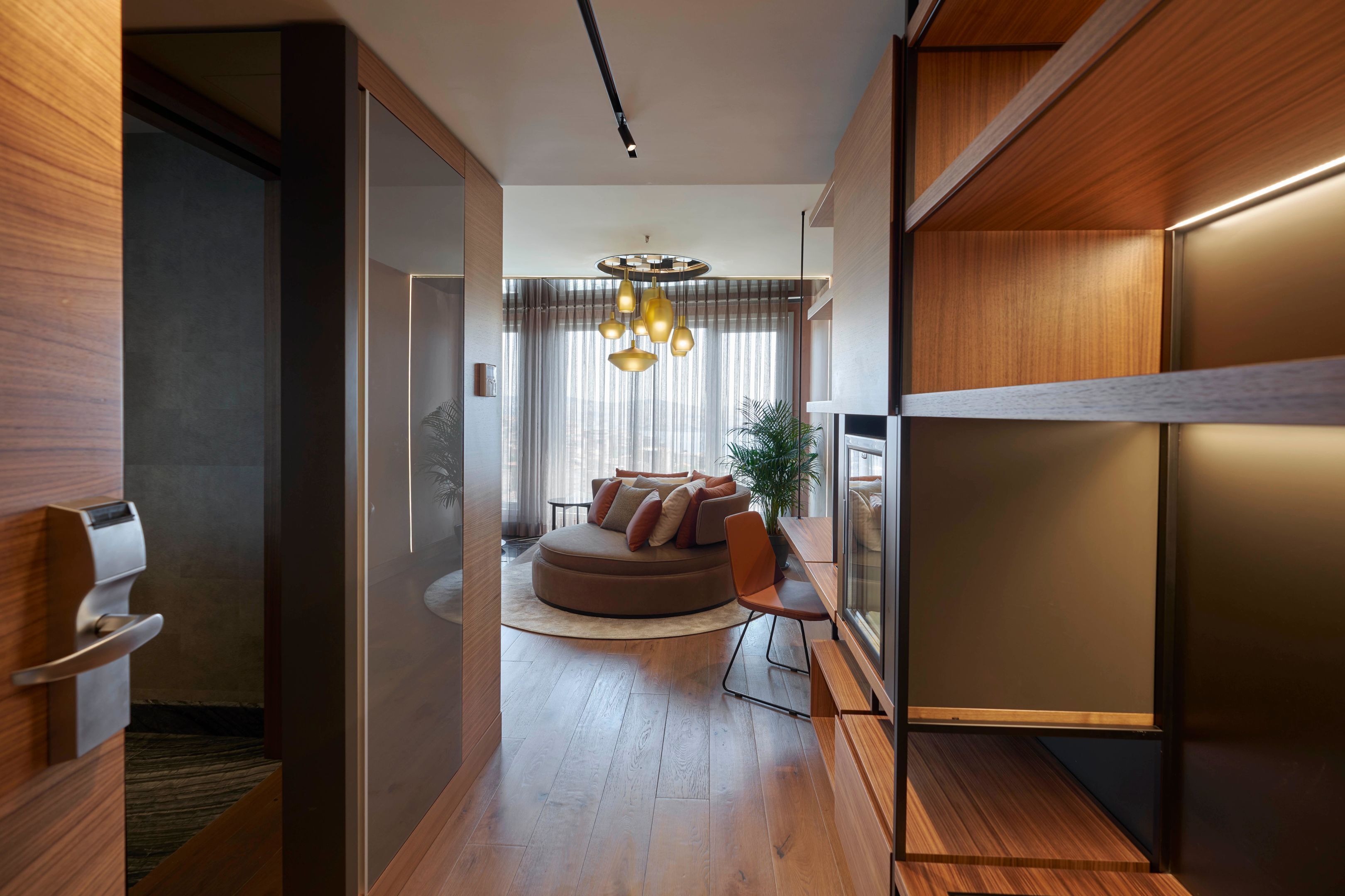 A modern interior with a wooden floor, a round seating area with cushions, a glass partition, and a hanging light fixture.