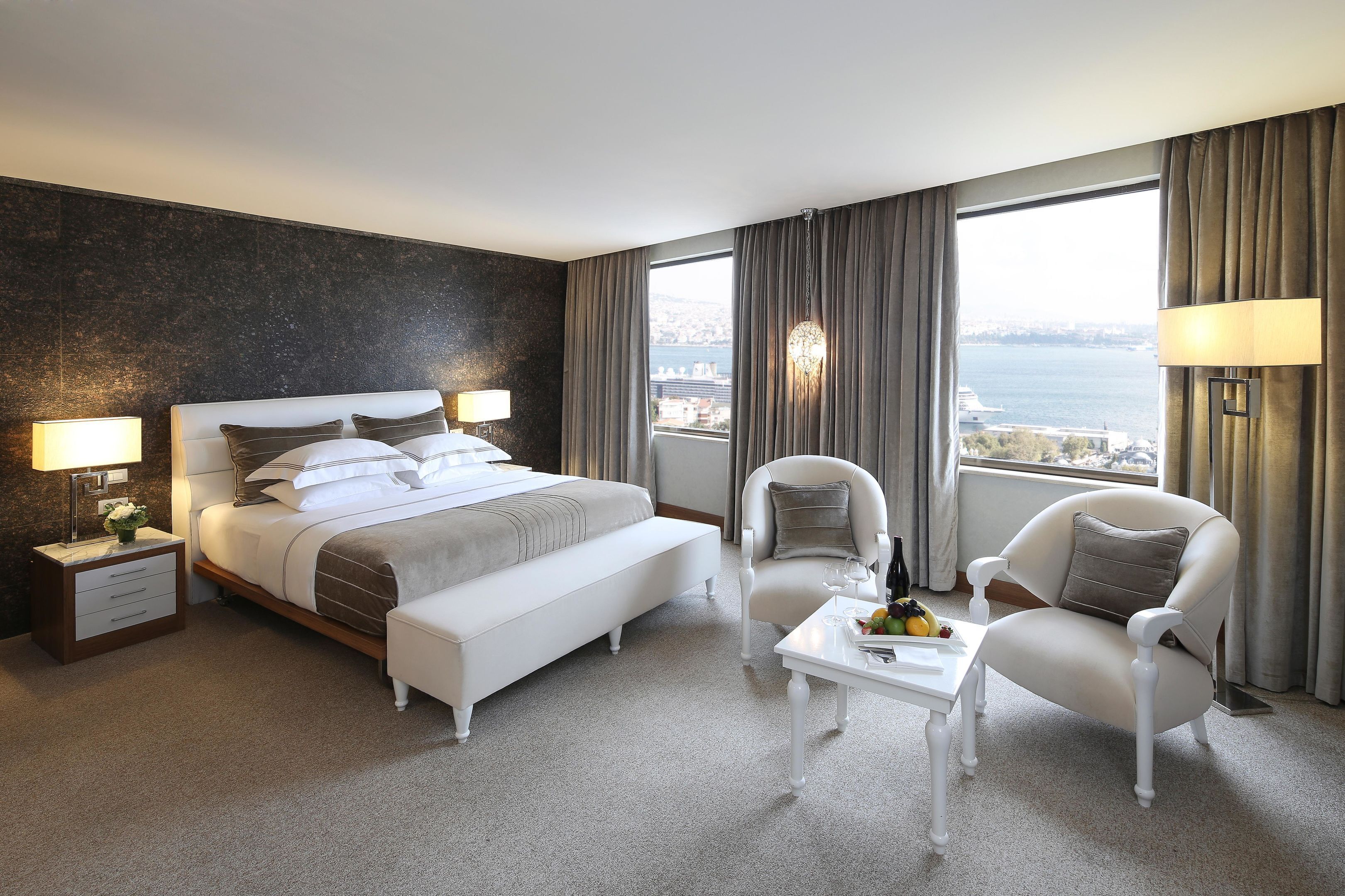 Une chambre d’hôtel avec un grand lit, une chaise, une petite table avec des fruits, et une vue sur la ville et l’eau.