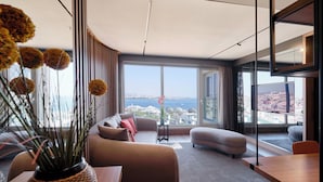 Een moderne woonkamer met een bank, voetenbankje en een eettafel. Er is een groot raam met uitzicht op de stad en een waterpartij.