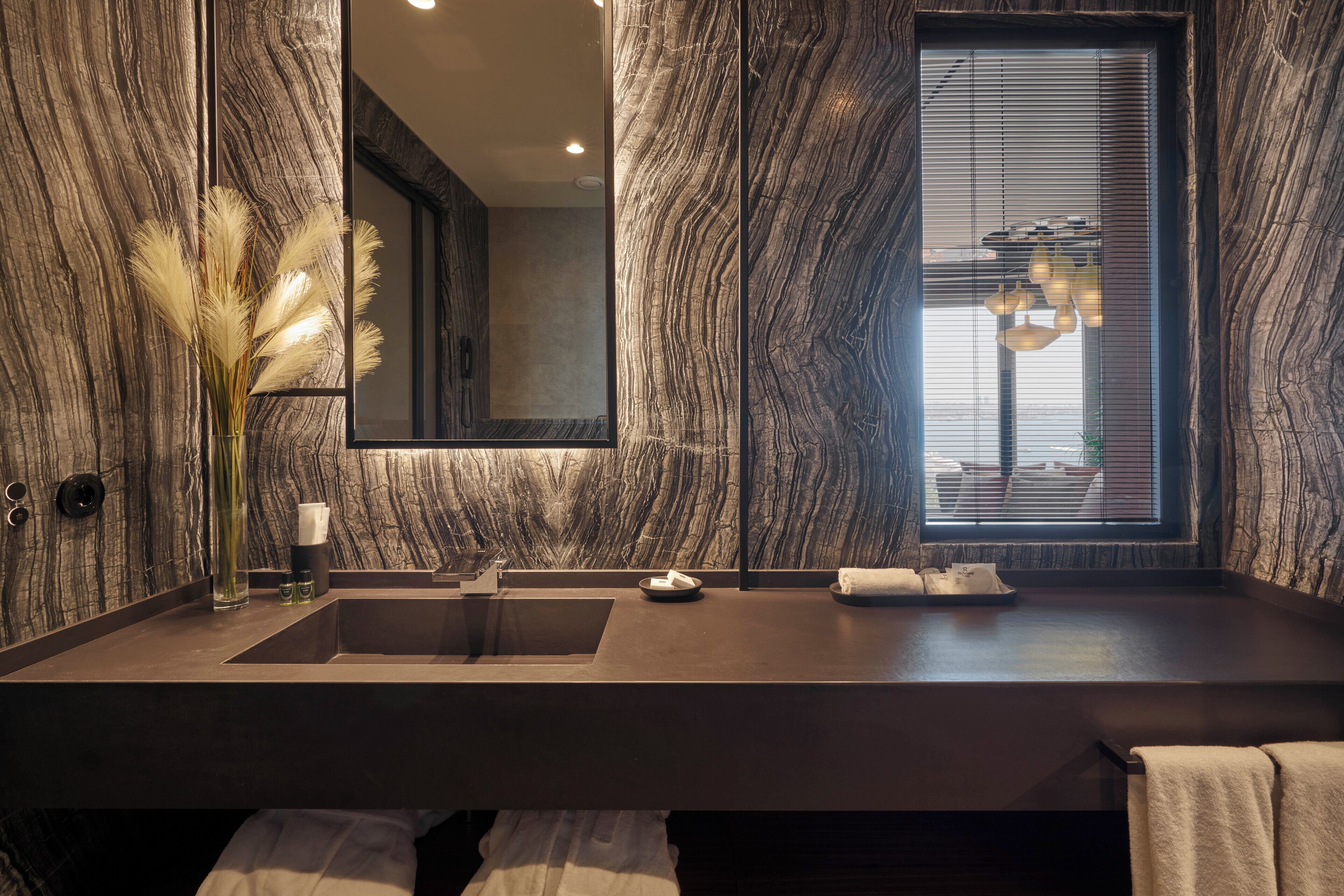 Une salle de bain moderne avec un mur en marbre, un grand miroir, un lavabo et une fenêtre avec des stores.