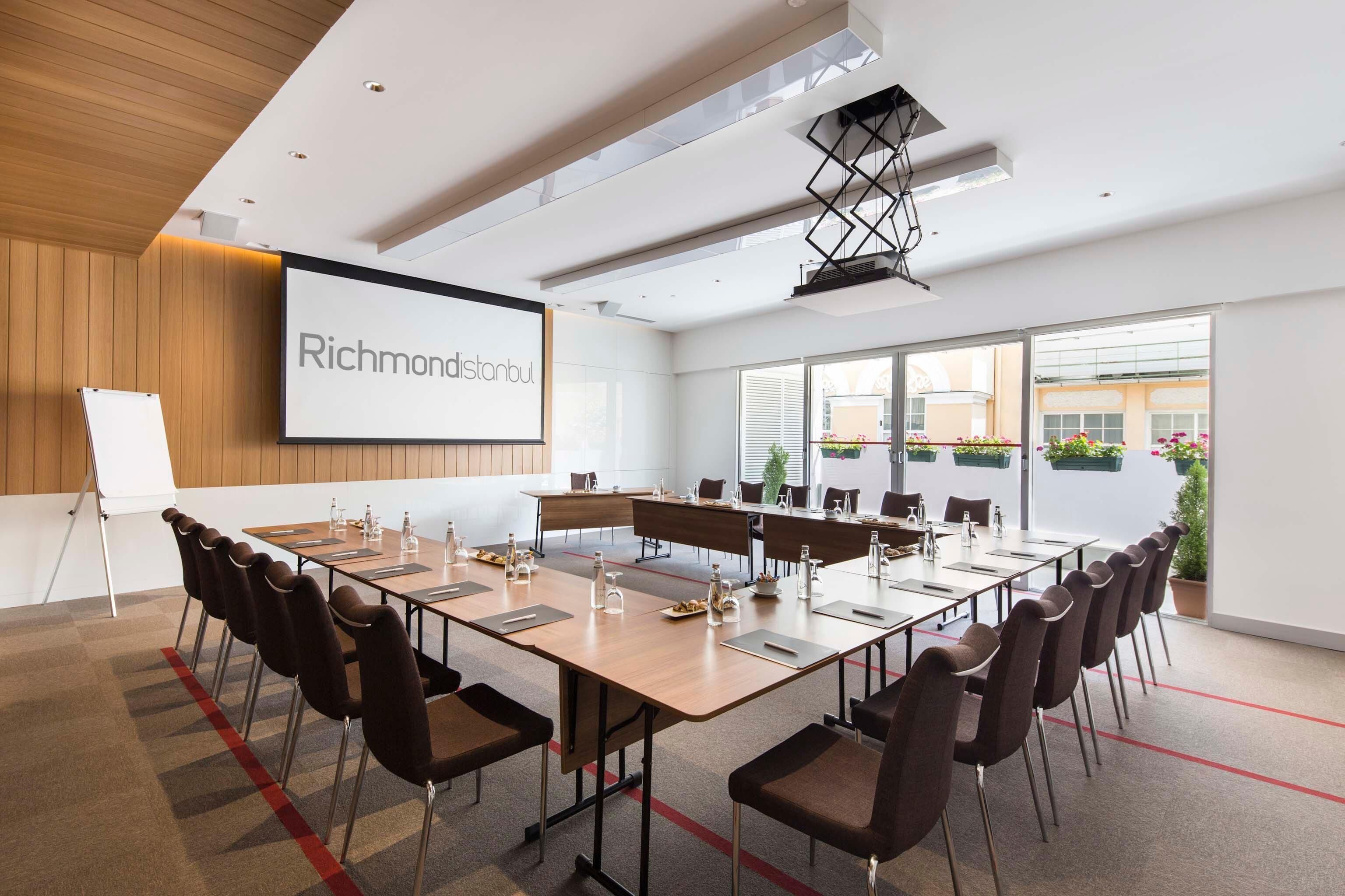 Ein Konferenzraum mit einer großen Leinwand, die „Richmond Istanbul“ zeigt, einem langen Tisch mit Stühlen und einer Whiteboard-Tafel.