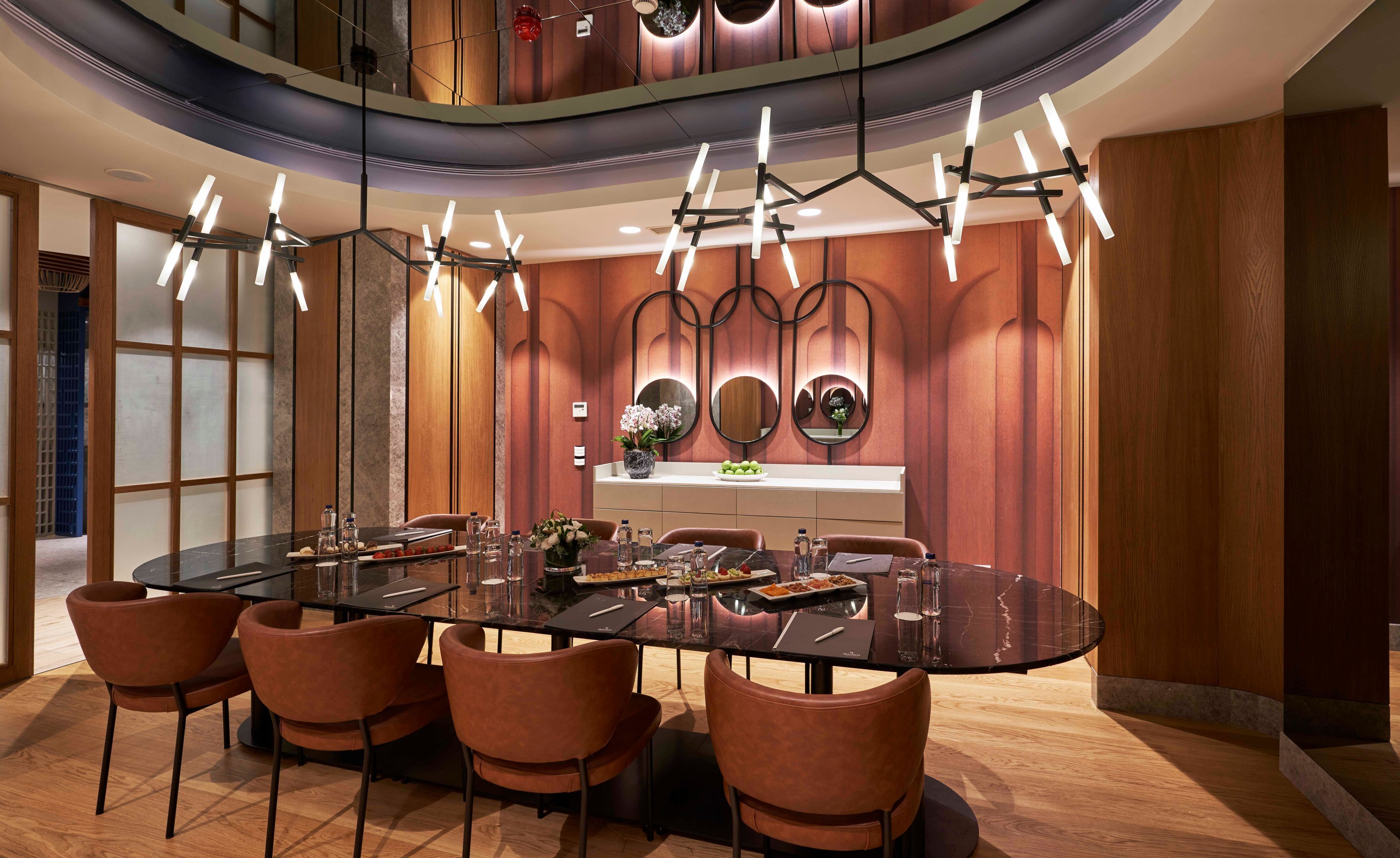 Een moderne eetruimte met een rond glazen tafel, zes bruine stoelen en een moderne kroonluchter.
