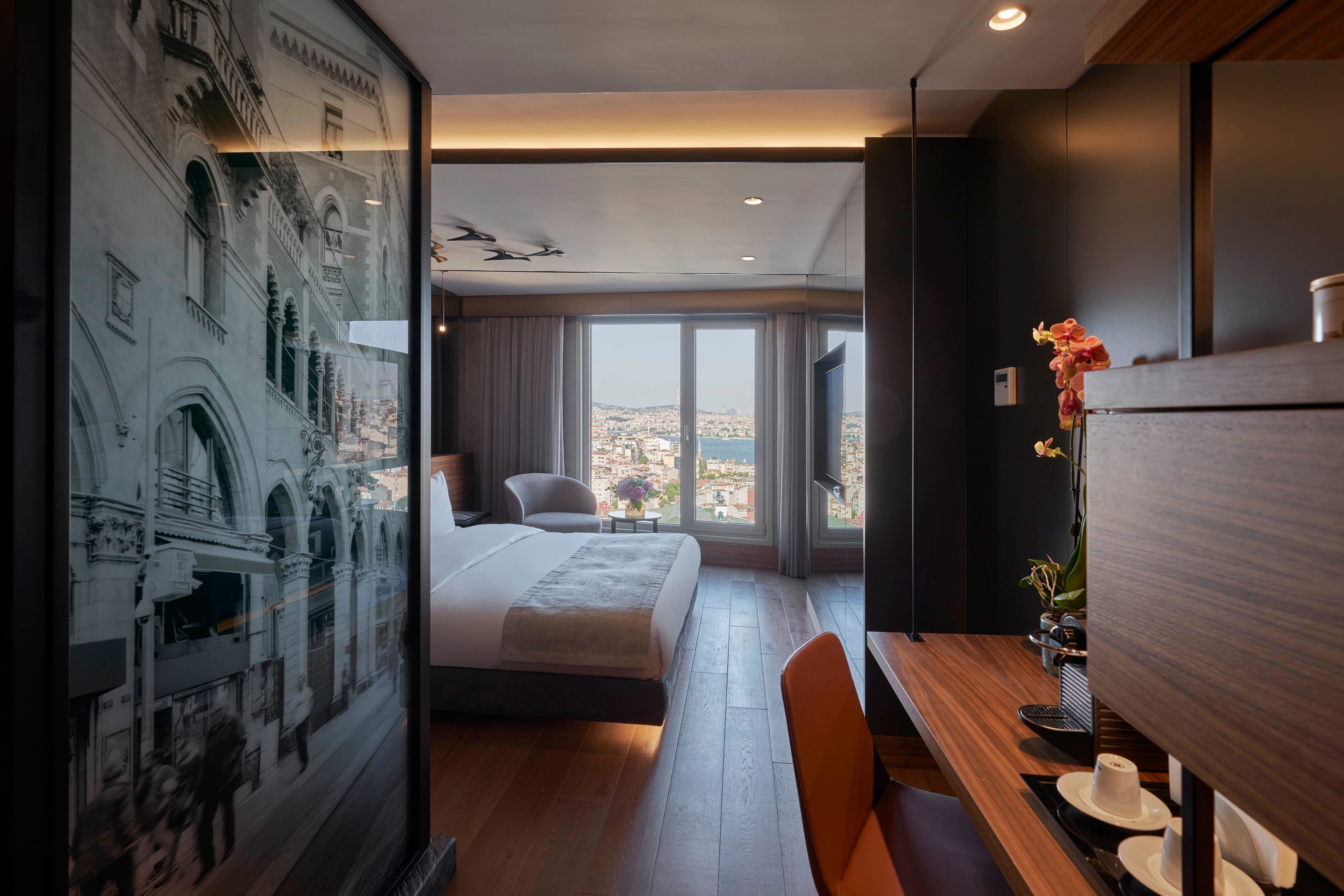 A modern hotel room with a large bed, a wooden desk, a chair, and a view of the city through large windows.