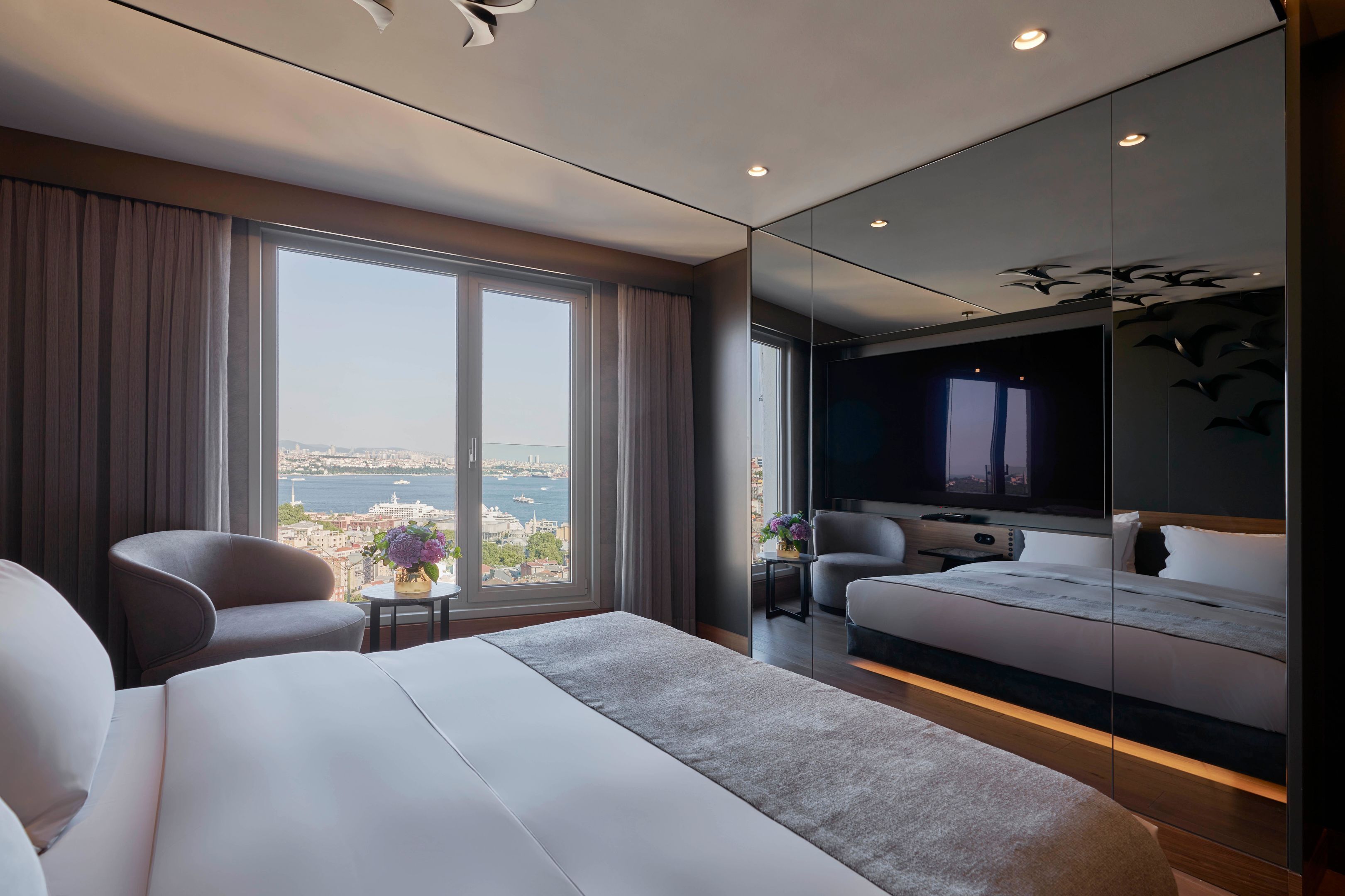 A modern hotel room with a large bed, a seating area with a small table, and a large window offering a city view.