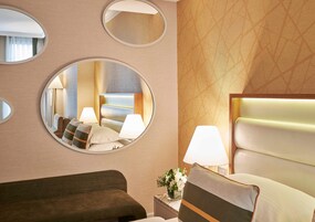 Habitación de hotel con cama, dos lámparas de noche, una mesita de noche con un florero y tres espejos en la pared.